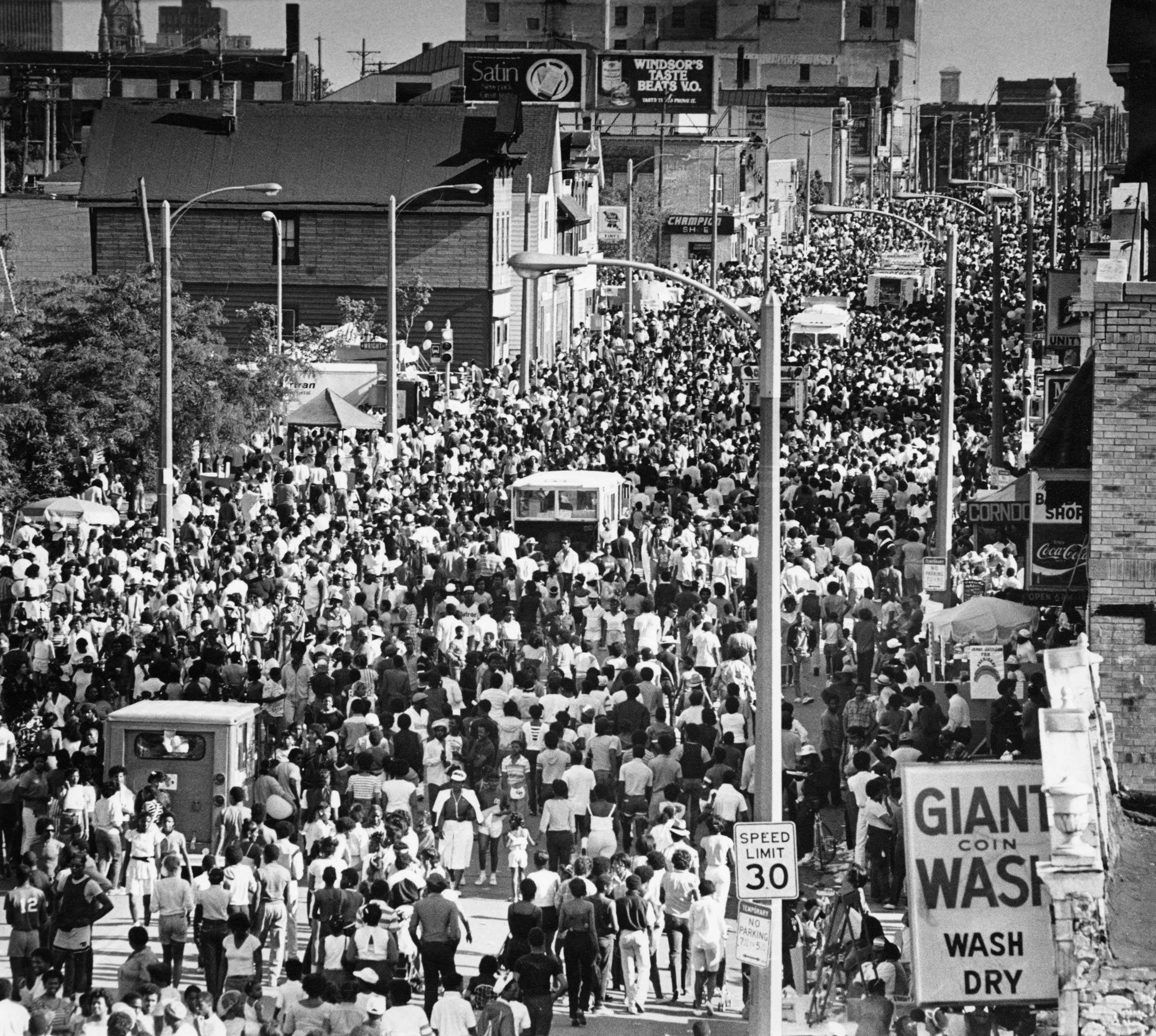 Black-and-white photo of a large crowd filling a city street, with storefronts and signs visible, suggesting a public event or gathering.