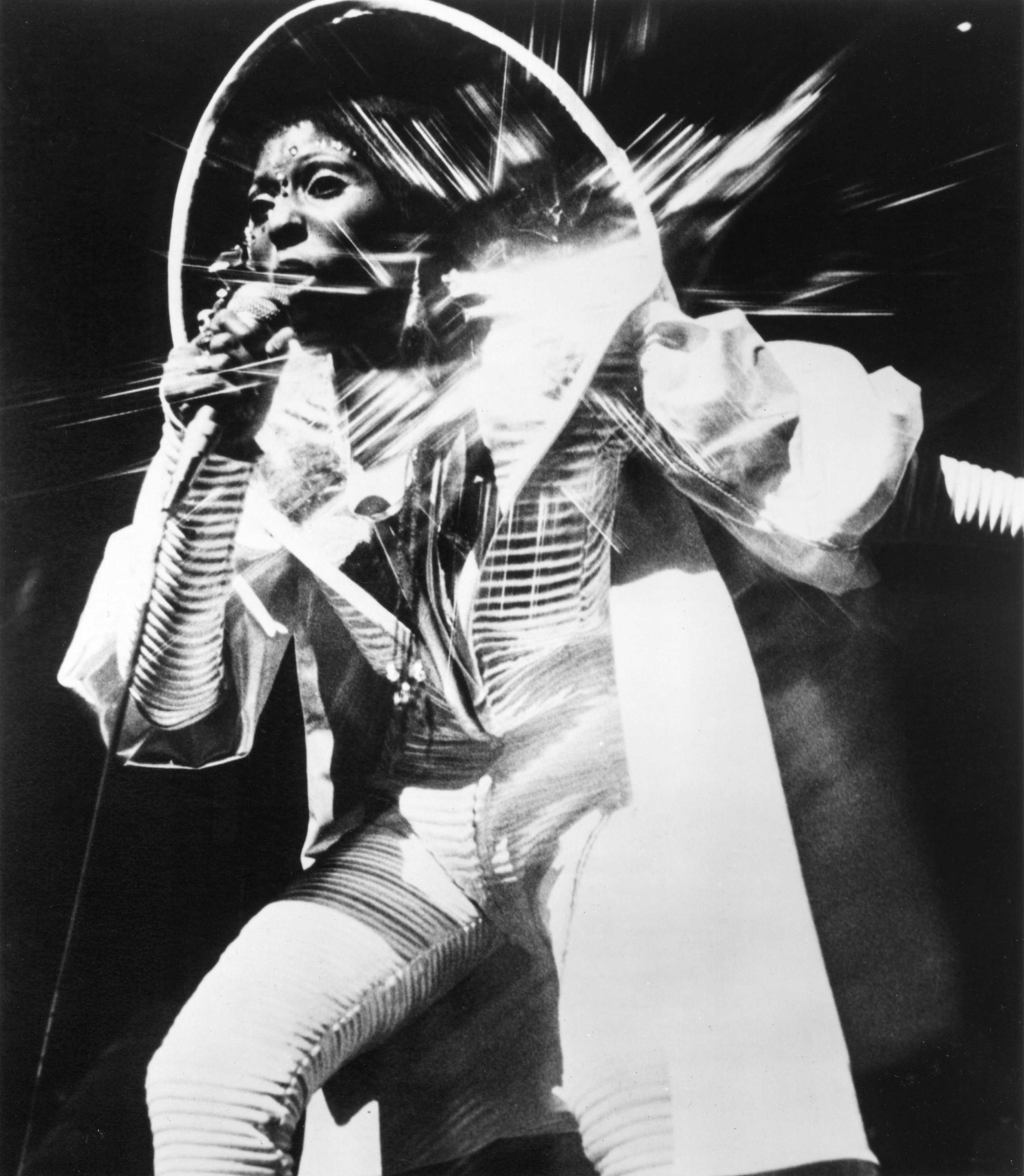 Nona Hendryx dressed in a white futuristic outfit singing into a microphone in this black and white photo.