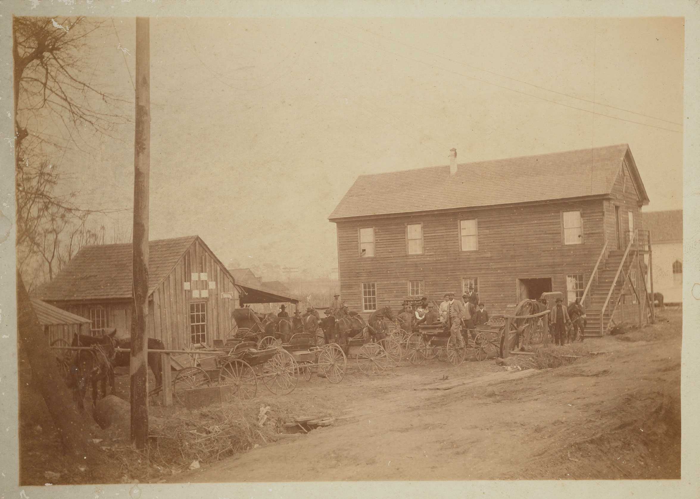 A worn sepia toned photograph of the horses and carriages outside of the blacksmith shop.