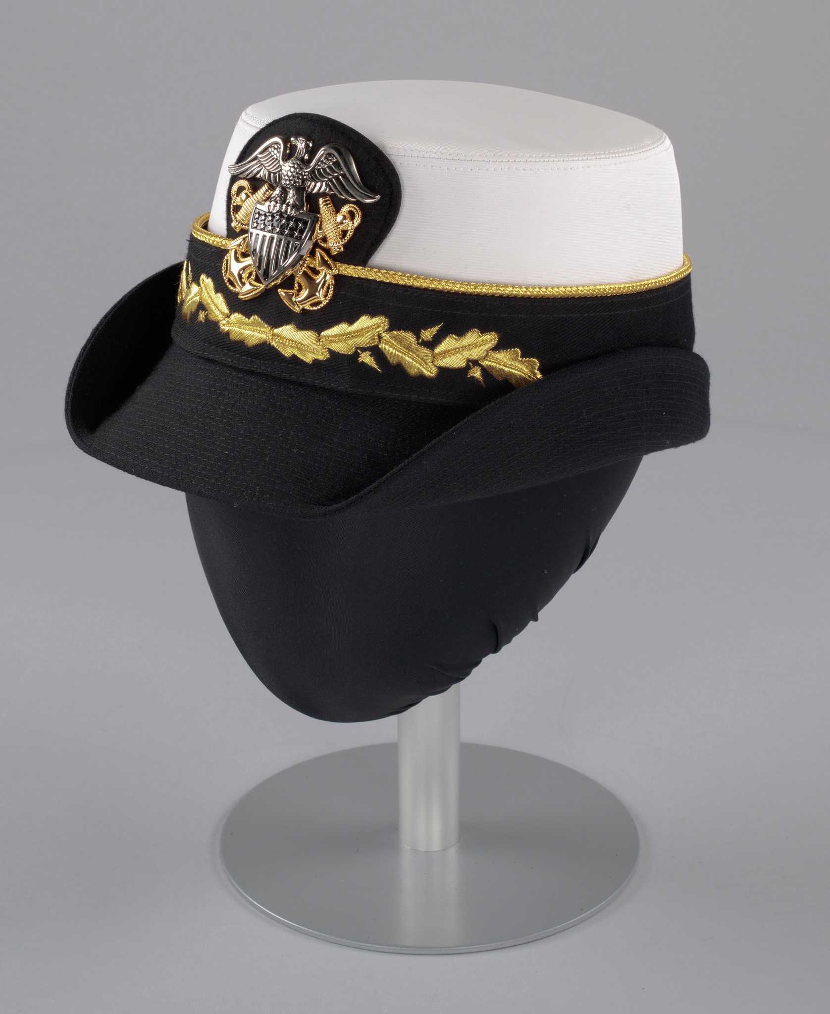 A US Navy dress uniform hat (a) worn by Admiral Michelle Howard as commander with a white waterproof fabric crown and a black canvas brim, with a hat band (b) featuring a US Navy insignia pin (c). The brim of the hat is stiffened, with both sides fixed in a turned up position. The interior of the hat is lined with black waterproof plastic with the manufacturer's name "Kingform Cap / De Luxe / NEW YORK" stamped on it in gold. The sides of the crown are lined in black synthetic satin fabric. There is a size "23" tag at the interior center back and a tag at the interior proper left back with style and military regulation information. 

The hat band (b) attaches to the hat (a) with four (4) metal snaps, one at the front and back and one on each side. The band is made from black canvas trimmed on the top edge with gold braid. A length of the black canvas cut on the bias is looped around the center back portion of the band and stitched to it at the bottom edge, with the tails left dangling below the brim of the hat (a). An oak leaf and acorn design is embroidered in metallic gold thread at the center front of the band. A piece of black felt backed in leather with rounded edges protrudes from the center front top edge of the band.

A large silver and gold metal US Navy insignia pin (c) with two screw back fasteners is attached to the black felt on the hat band (b). The eagle and shield are in silver, while the two crossed anchors are in gold.