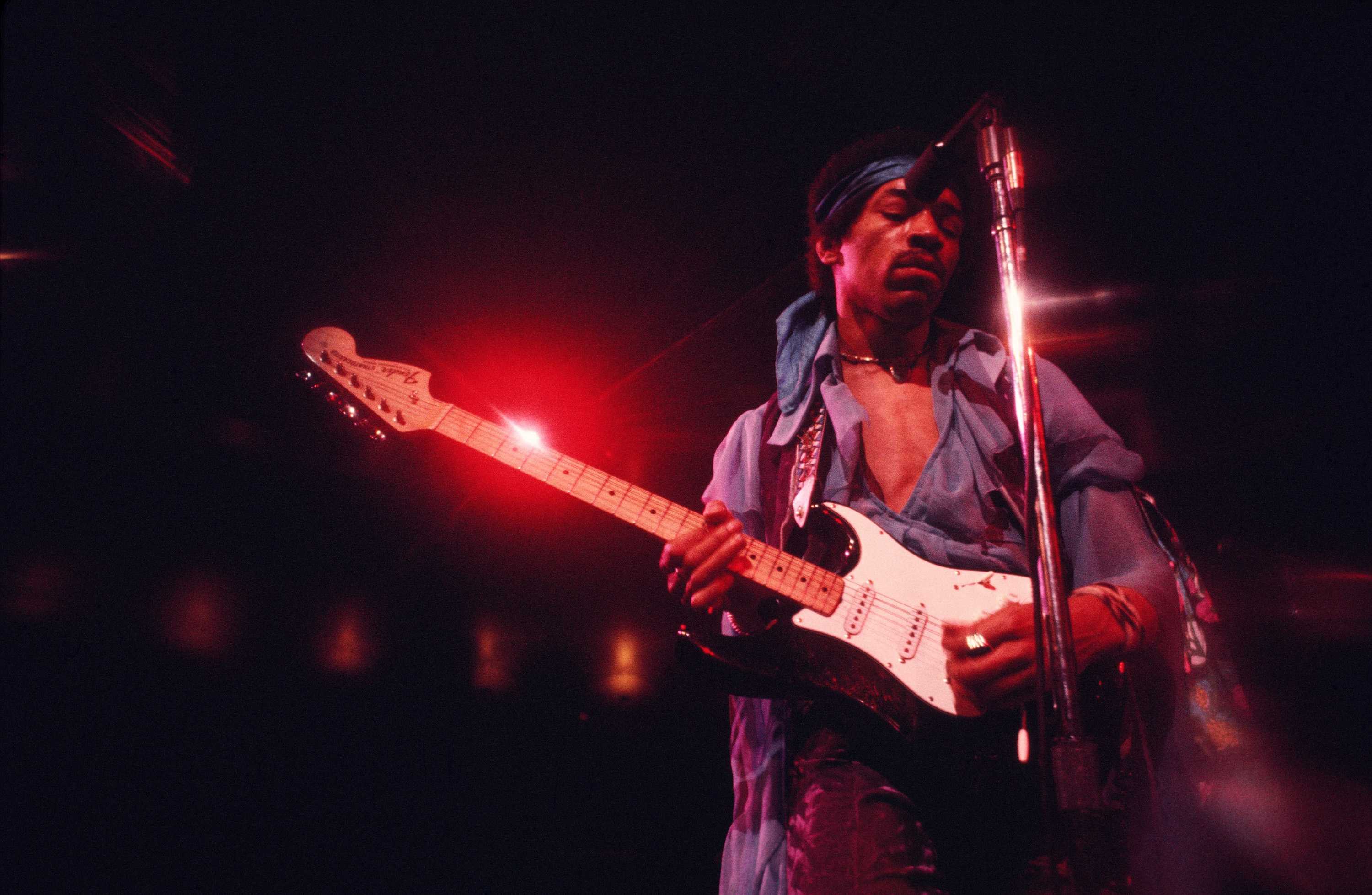 Jimi Hendrix is playing a black and white electric guitar in a blue suit, loosly button to expose his chest.