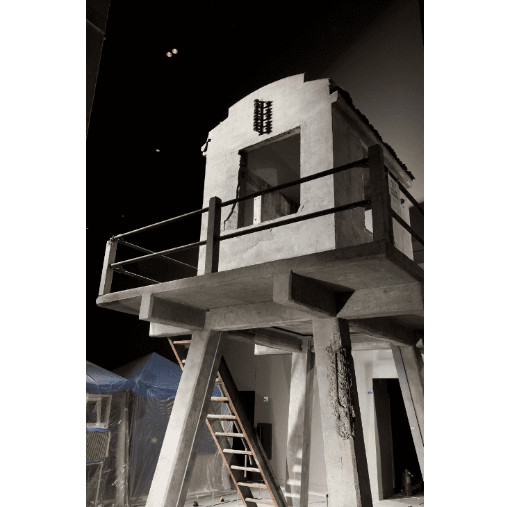 A concrete guard tower with a ladder leading up to a platform and a small structure for guards to stand inside.