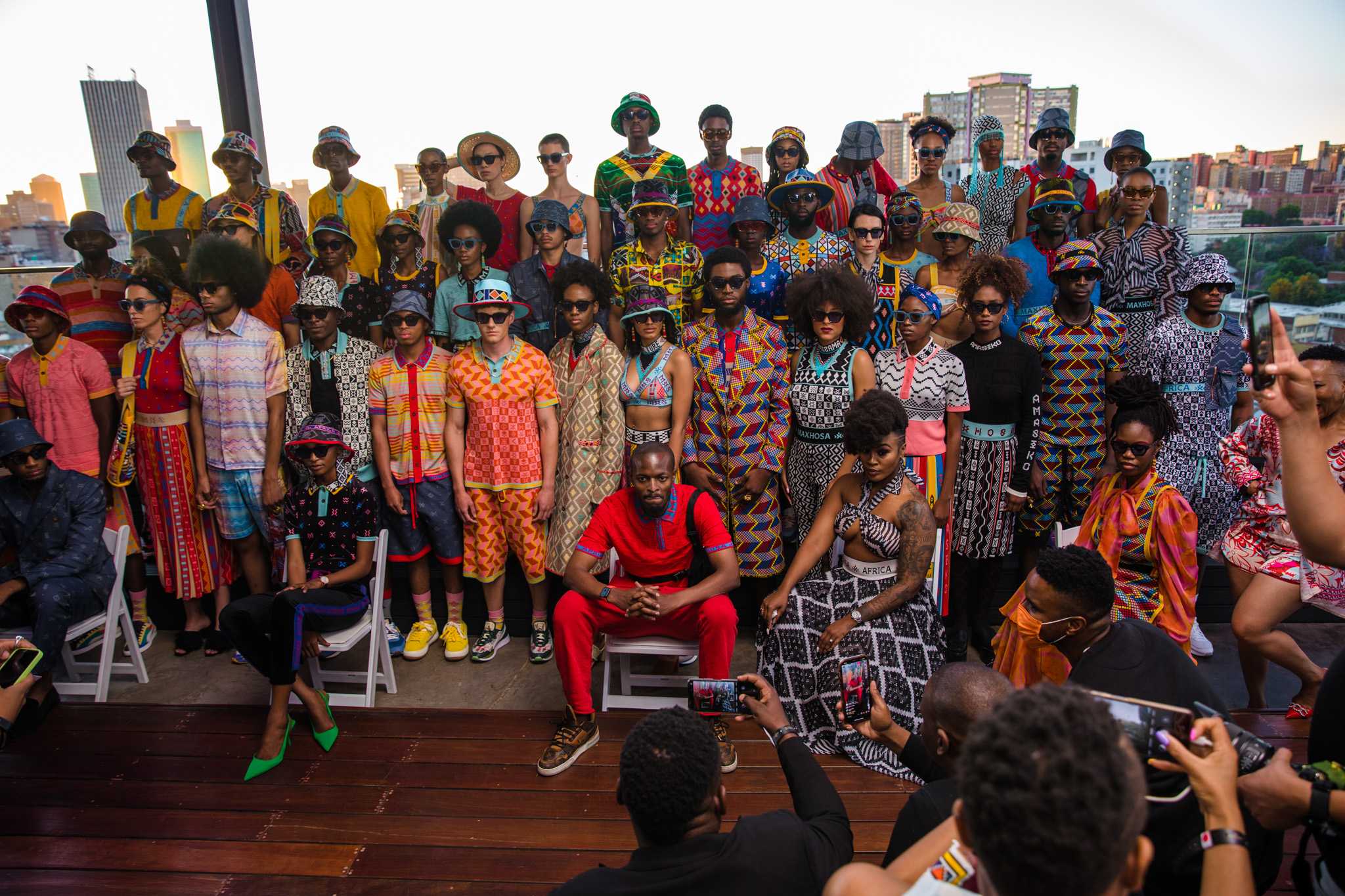 Models and fashion designing standing and sitting on a dark wood stage in his colorful collection.