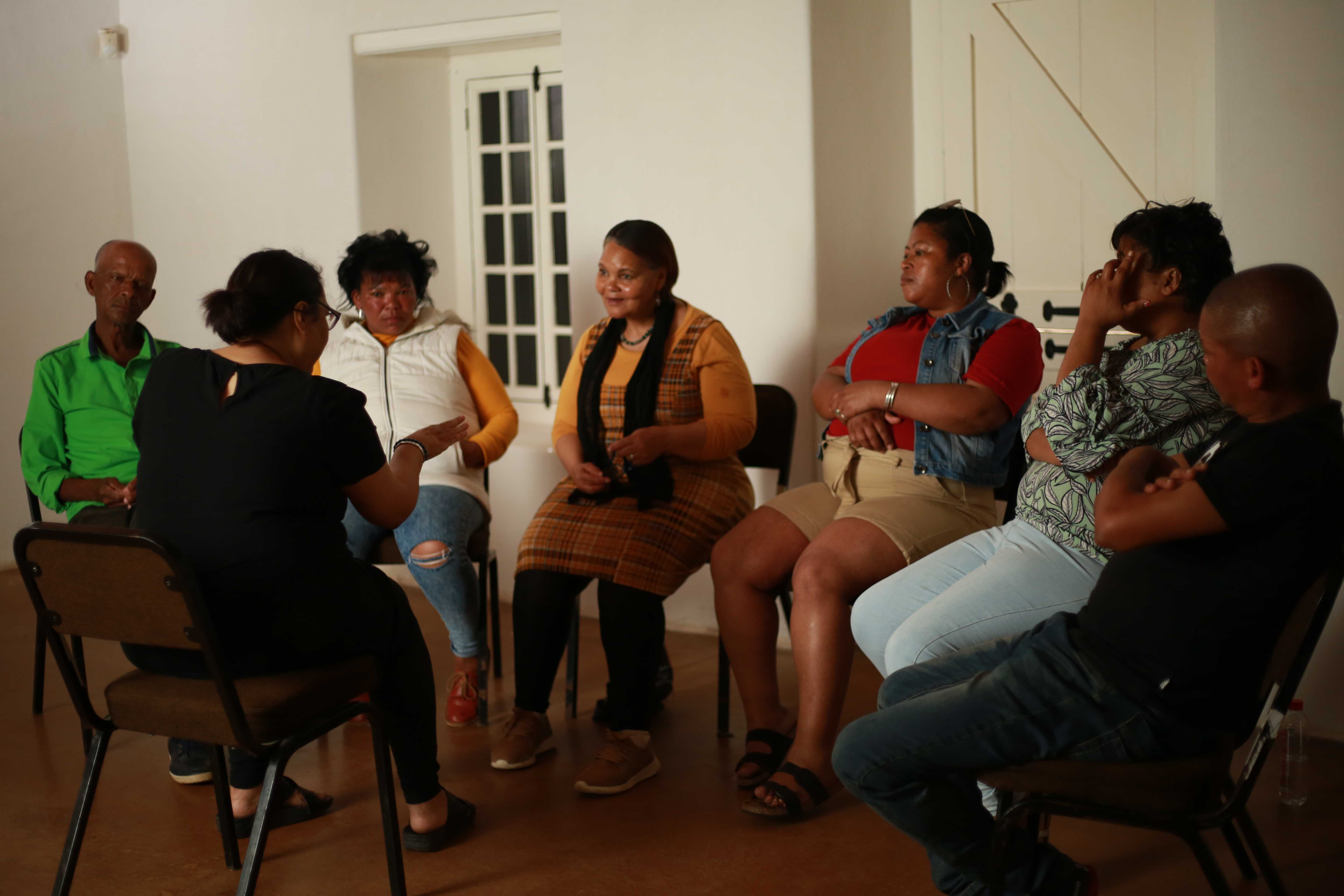 A small group of people seated in a circle of chairs during a discussion or meeting in a bright indoor room.