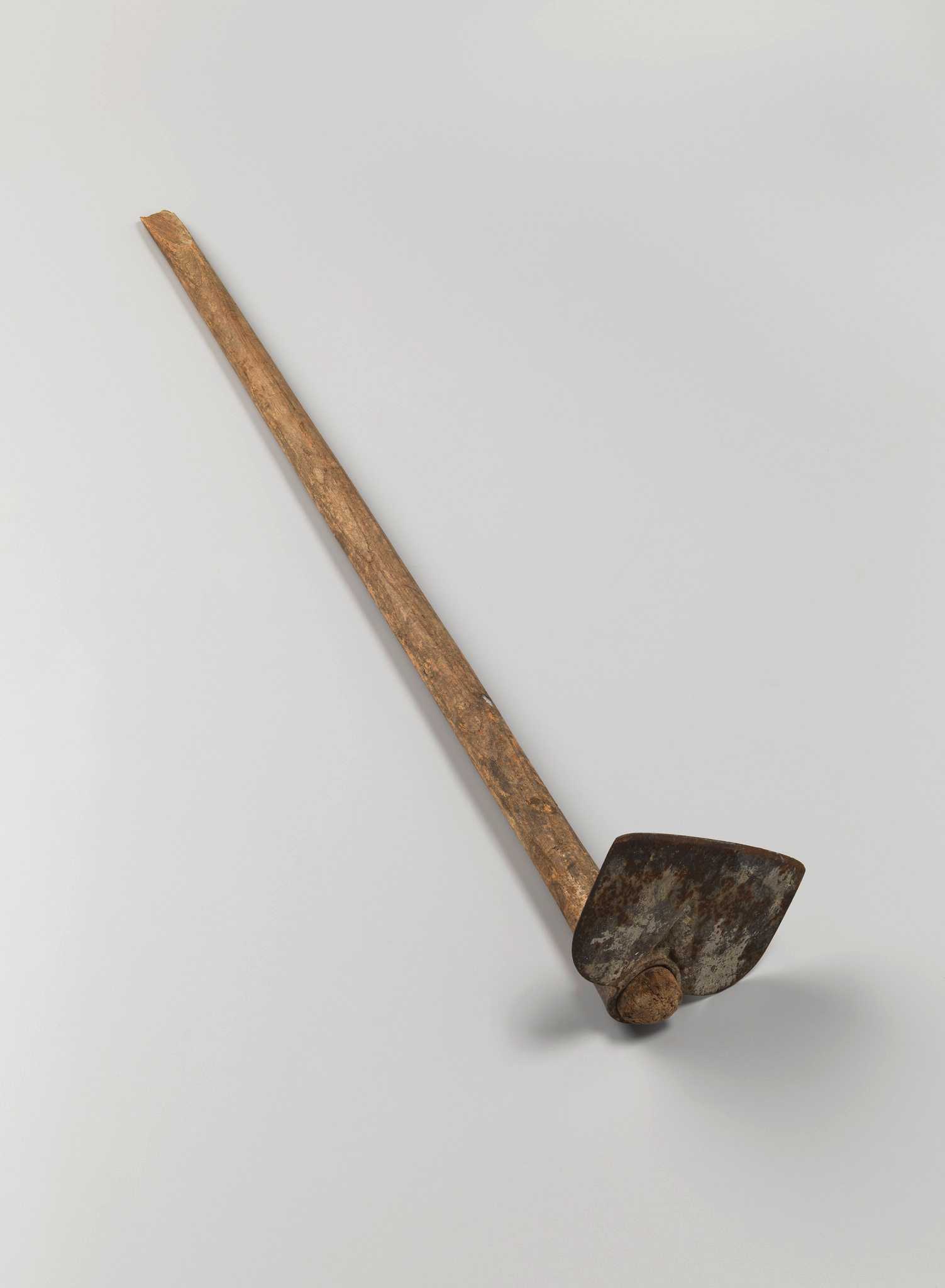 A draw hoe with a wooden handle. The hoe is mounted perpendicularly to the handle through an eye at the base of the hoe head. The hoe blade has rounded shoulders and a straight cutting edge. The handle is long and rounded. Near the top of the handle, the wood is split diagonally.