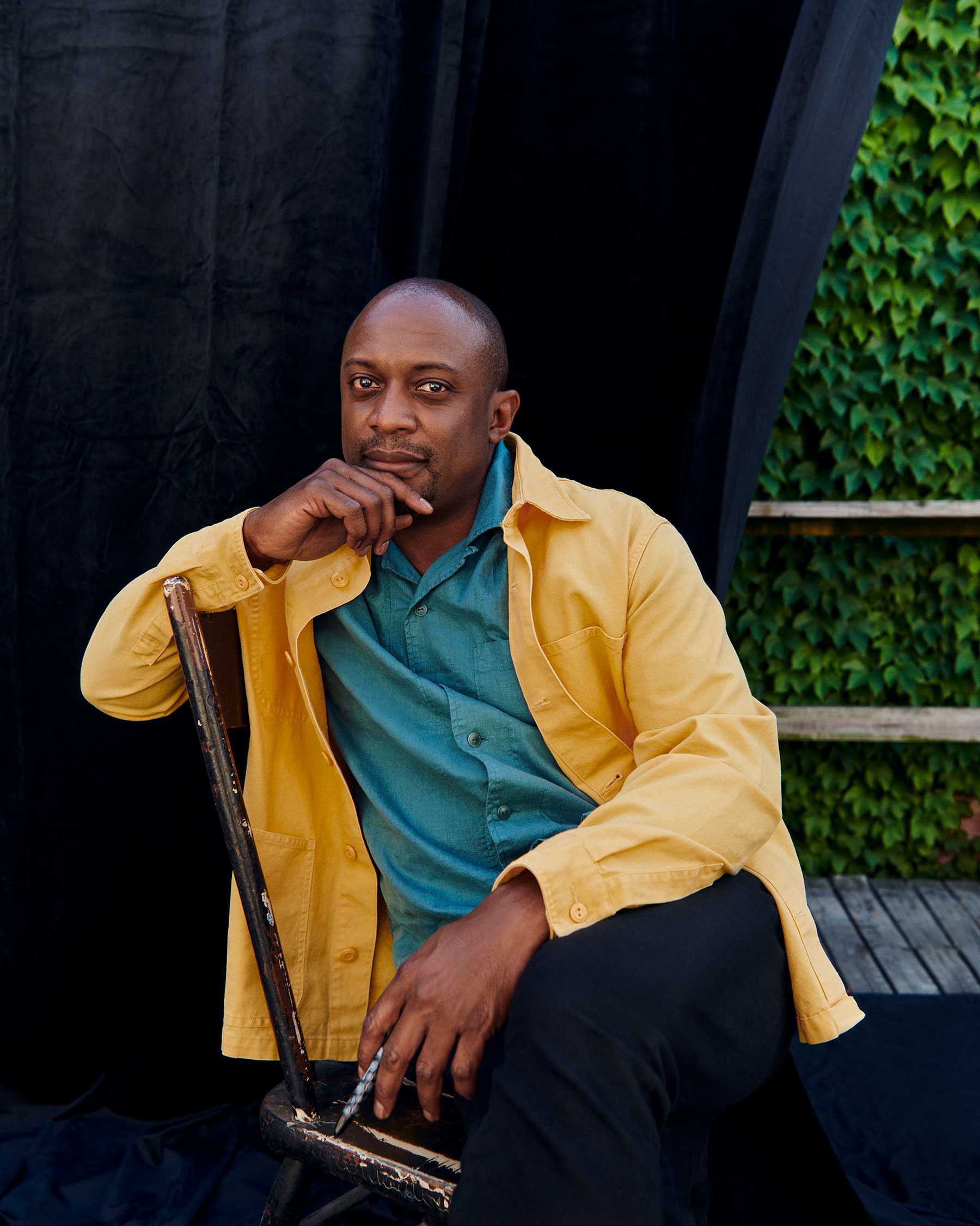 Man seated on a chair with a contemplative expression, wearing a yellow jacket over a blue shirt, set against a black curtain backdrop.
