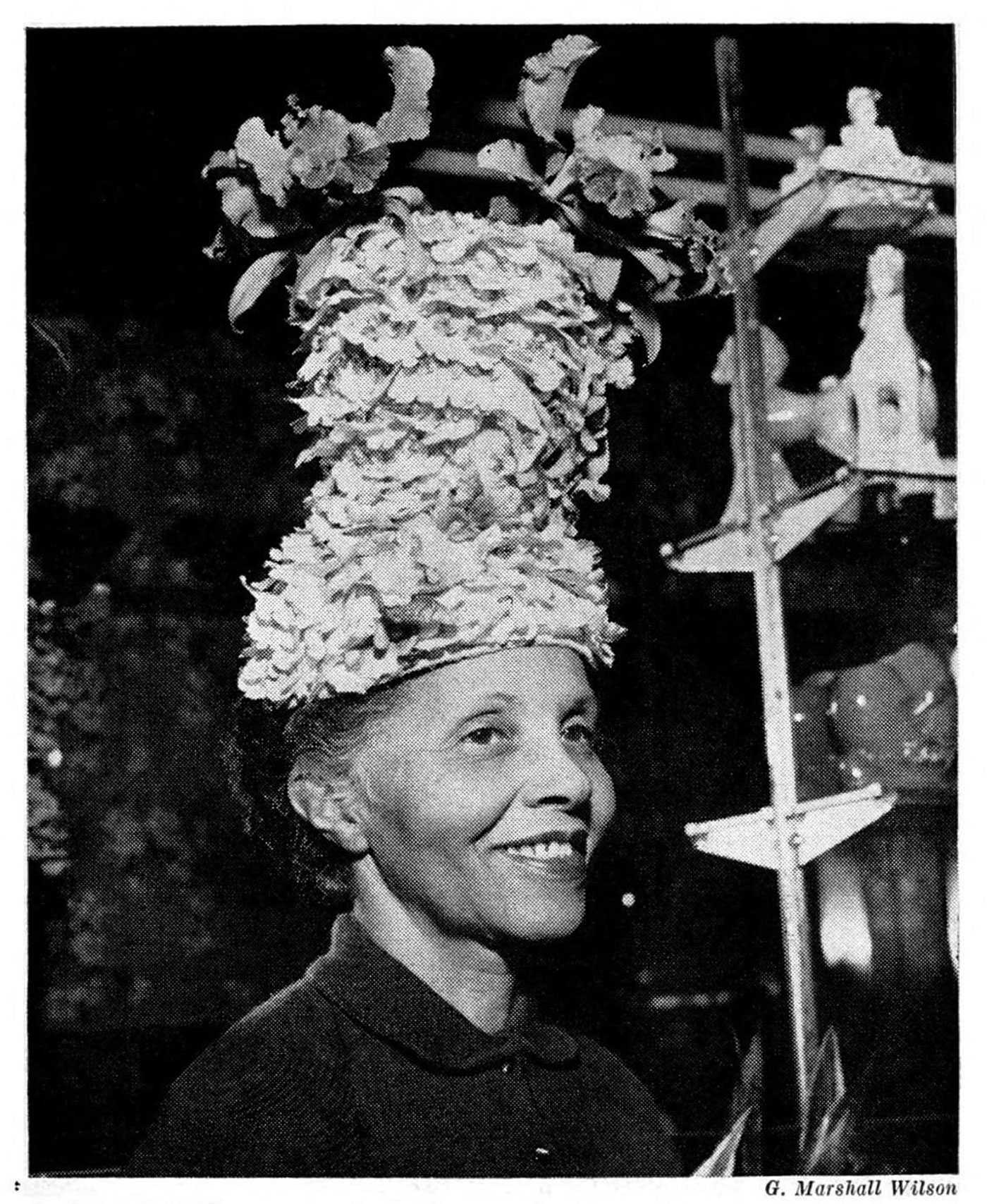 A woman wearing an elaborate floral hat with a rack of hats in the background.