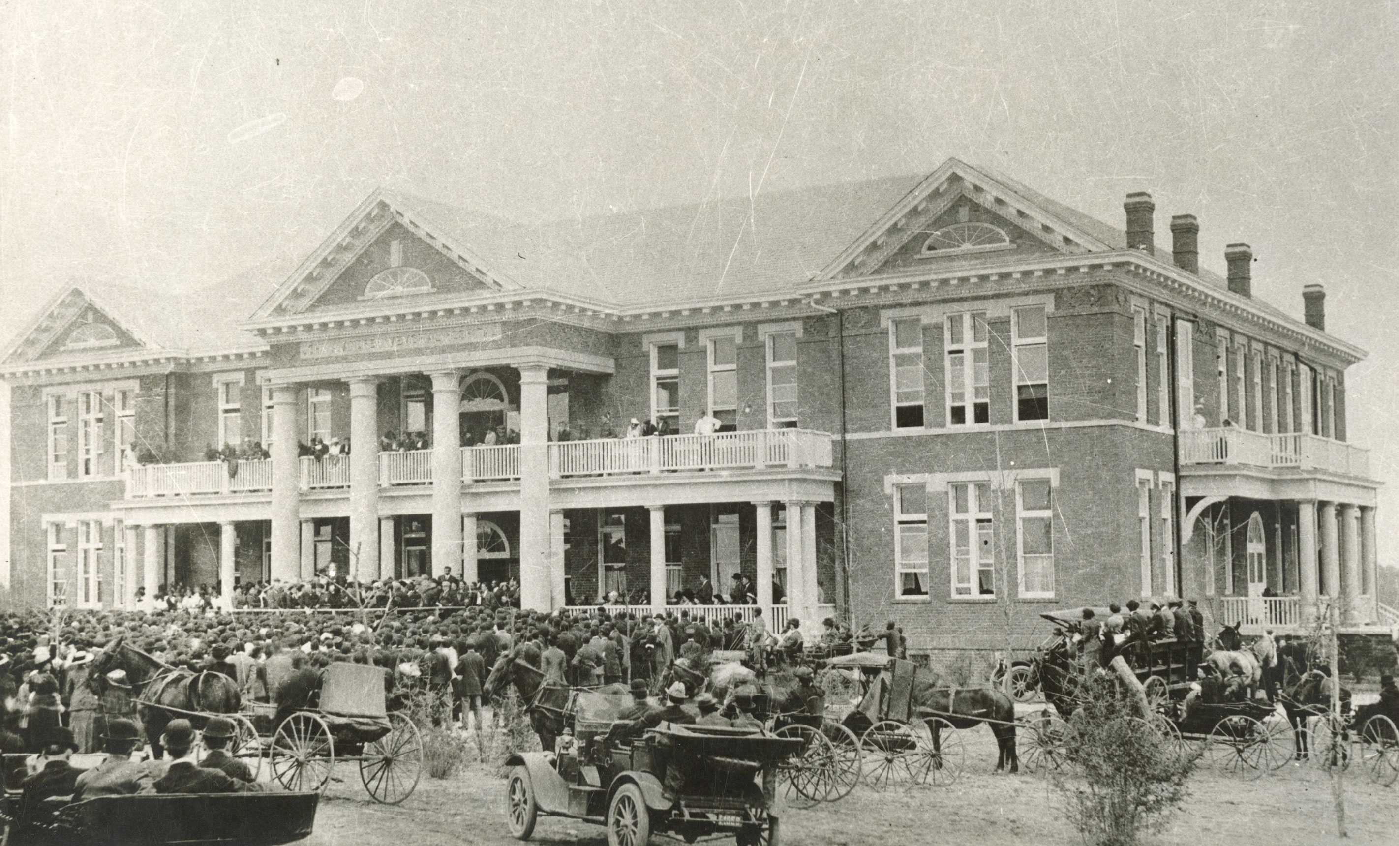 Photo of a large brick building with a crowd gathered and horse-drawn carriages in the foreground.