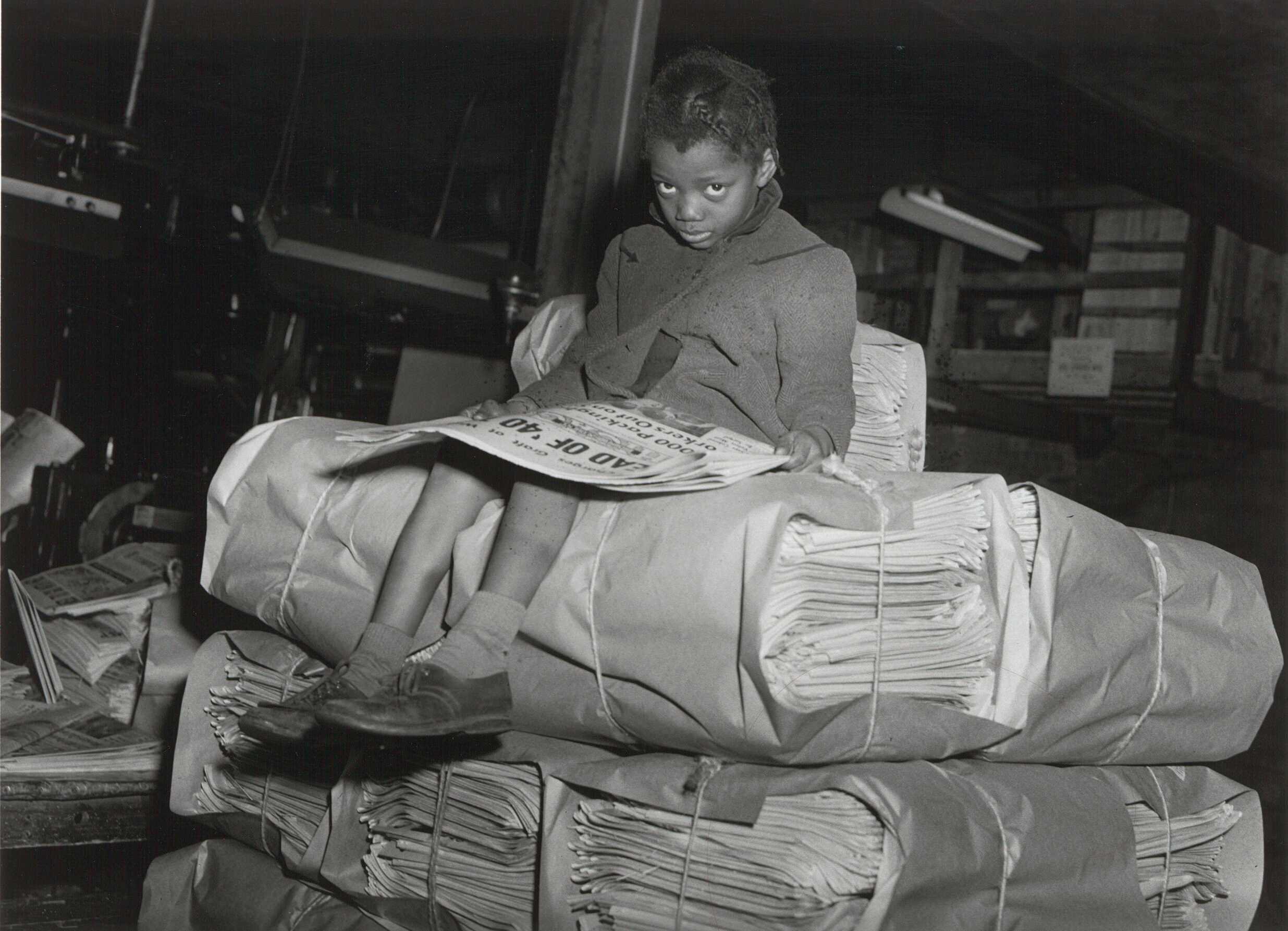 A black and white photograph of a young girl sitting on a pallet of Pittsburgh Courier newspapers.