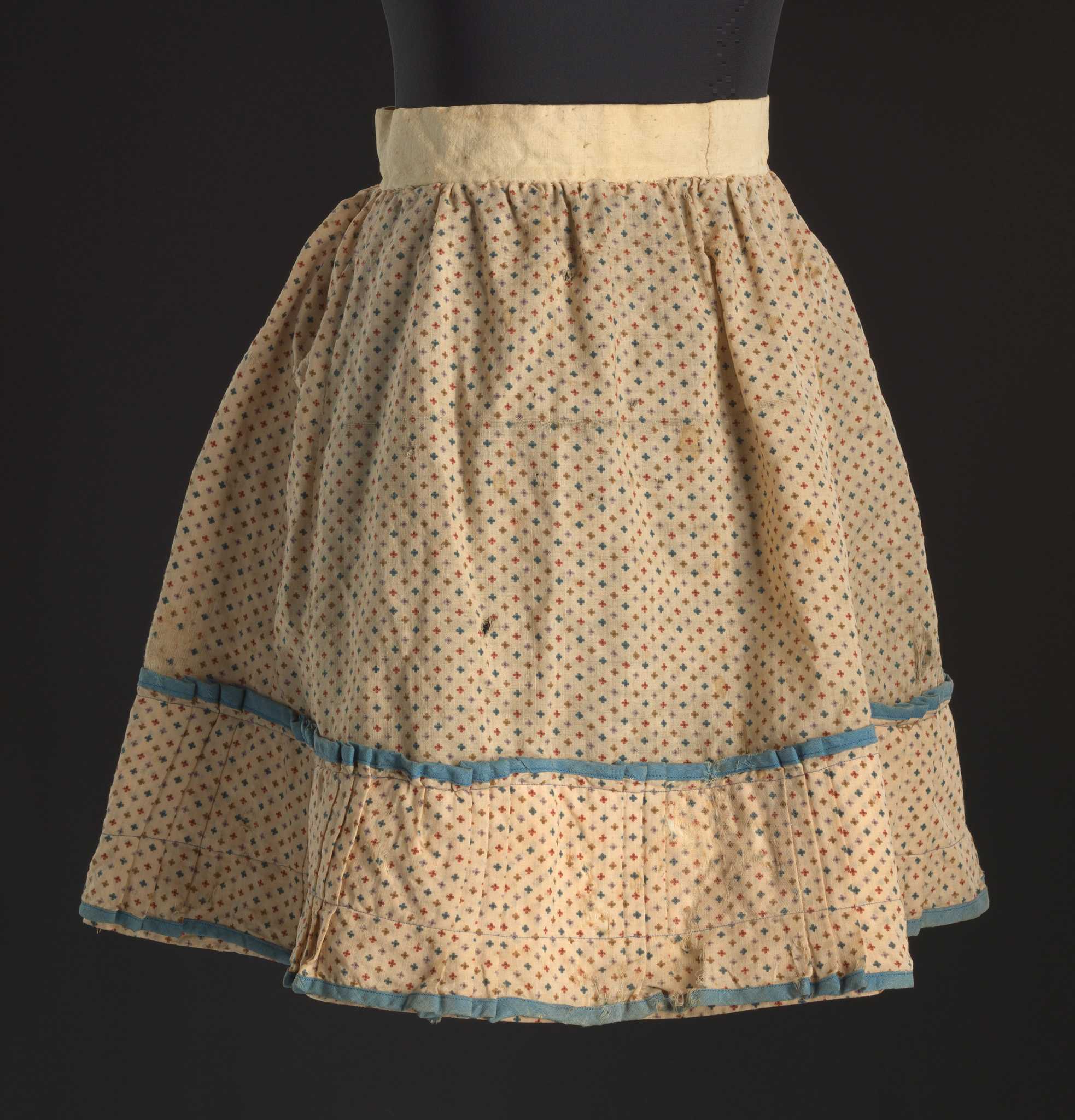 A child's circular skirt in a floral print with a pleated flounce on the hem. The main fabric is a plain weave natural fiber fabric with a cream ground and a repeating pattern of small four-petal flowers in red, purple, blue, and tan. The skirt is predominantly hand-sewn. It has a waistband made from an undyed bast fiber fabric with the floral print gathered and whip stitched to the waistband. The skirt is more closely gathered on the back. There is a slit at the center back that divides the waistband completely. There is no attached closure method, but rust staining at the back opening of the waistband suggests the skirt was pinned to close. The bottom of the skirt has an extra flounce of the floral print that has a series of pleats around the skirt in sets of three alternating with straight sections. This flounce is machine-sewn at the top and 1 1/2 inches from the bottom using blue thread that is possibly a synthetic fiber. The bottom and top edges of the flounce are bound with blue fabric sewn on the bias. The interior of the skirt is lined only behind the extra flounce with a hand-pinked undyed bast fiber fabric. There is a previous repair on the back proper right side of the skirt where a panel of light brown synthetic fabric is hand sewn on the interior of the skirt from the gathering at the waist to the top of the older linen lining on the bottom.
