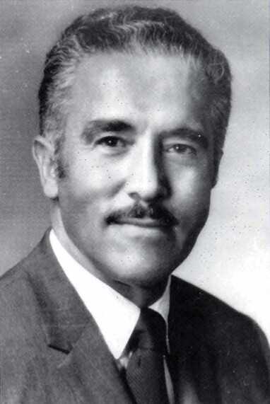 Black-and-white portrait of a person wearing a suit, white shirt, and dark tie against a plain background.