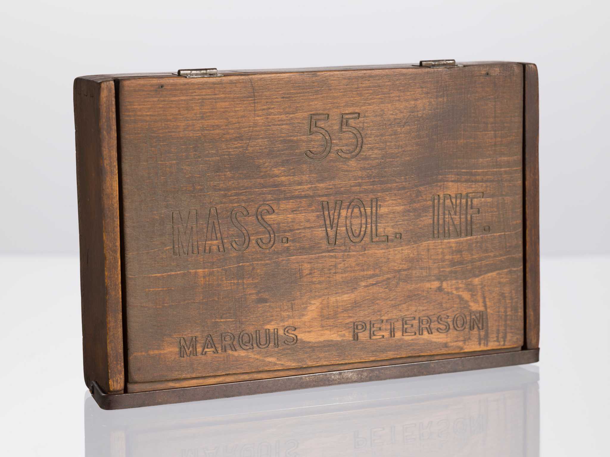 A wooden box owned by Marquis Peterson. The rectangular box is varnished brown and has a lid on top. Embossed on the lid is “55 / Mass. Vol. Inf. / Marquis Peterson.” The lid is connected to the box by two metal hinges. The sides of the box are constructed with dovetail joints and the bottom is nailed in place. A metal clasp runs the length of the box and secures the lid in place at front.