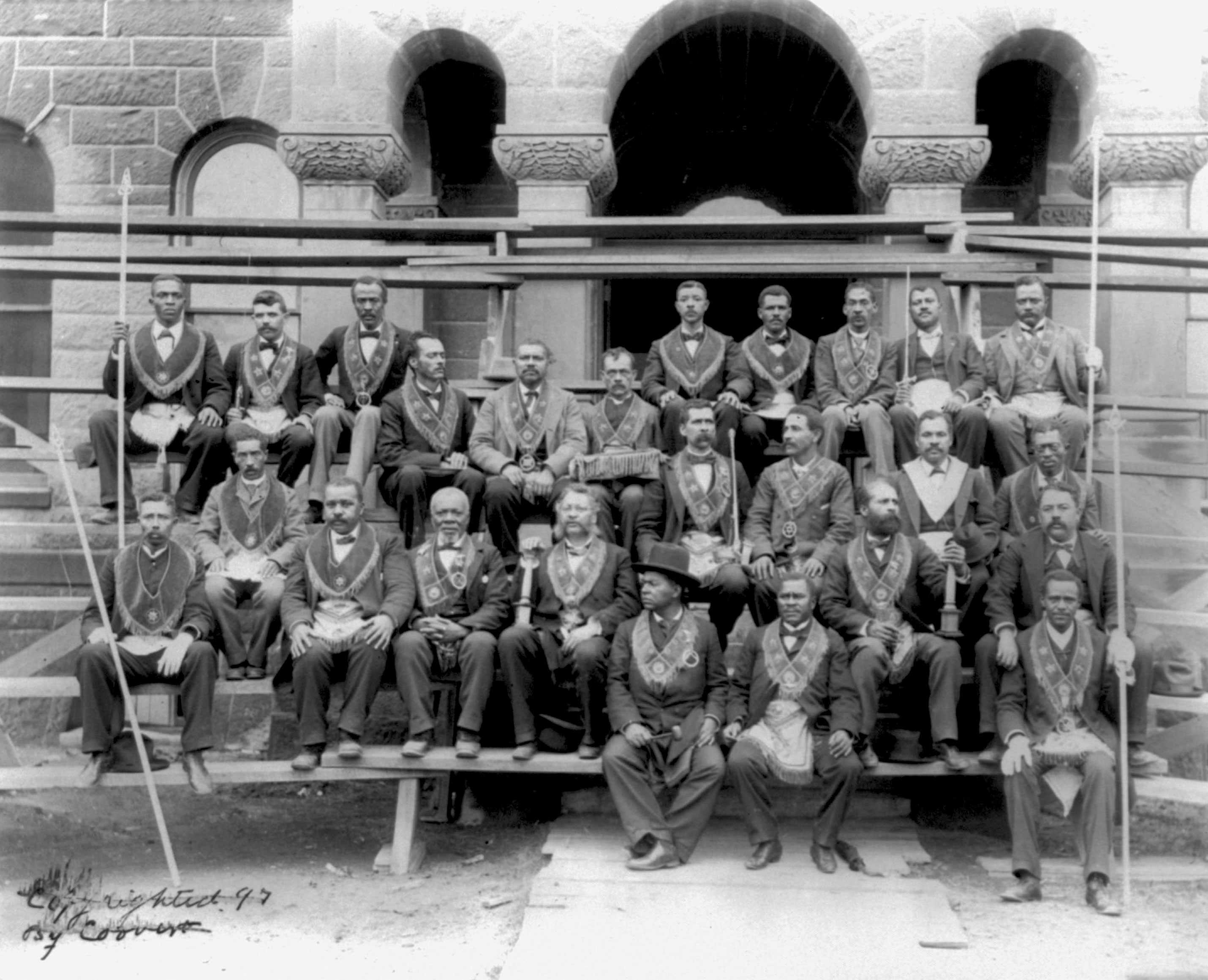 A group of 15 or so Prince Hall Masons sit on bleachers, posing for a photo.