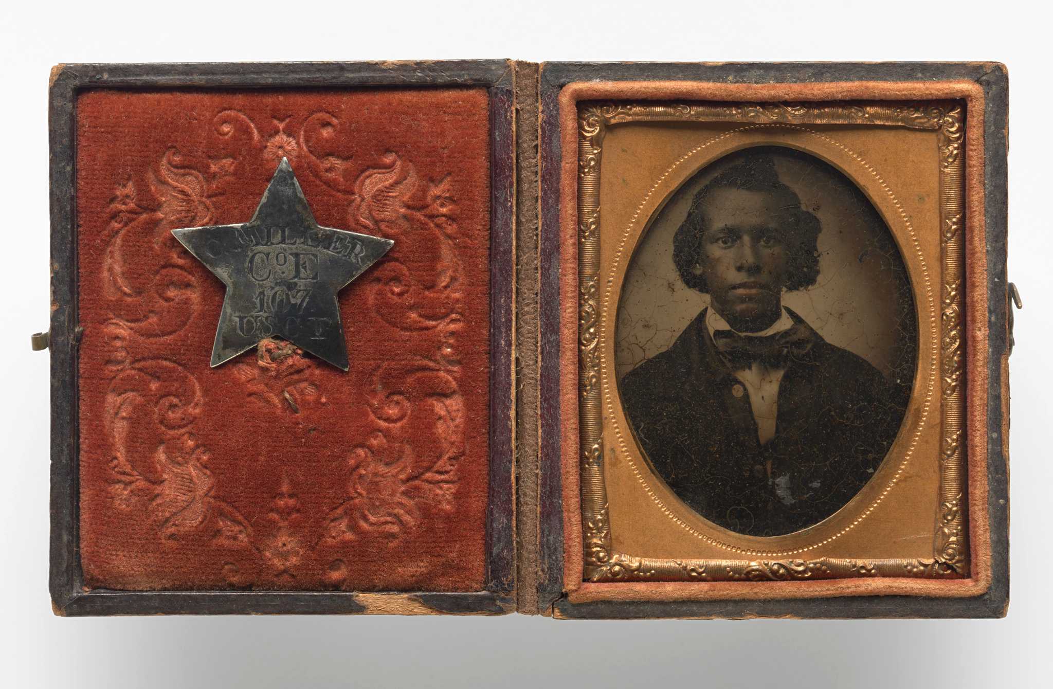 Tintype of Creed Miller, a soldier of the Kentucky 107th Regiment, Company E (and later Company C), United States Colored Troops. The tintype is encased in a copper scrolled frame with an oval window within a red velvet-lined, brown leather box. The tintype depicts Creed from the waist up in a dark overcoat, white collared shirt and bow-tie. His hair is styled and parted to the side. His miltary identification pin is fastened to the red velvet fabric on the interior of the case. The pin is a silver five pointed star with  [C. MILLER / Co. E / 107 / USCT] engraved at center. The front and back of the case is brown leather with detailed scroll work consisting of vines and flowers. The case has a metal hook and eye closure.
