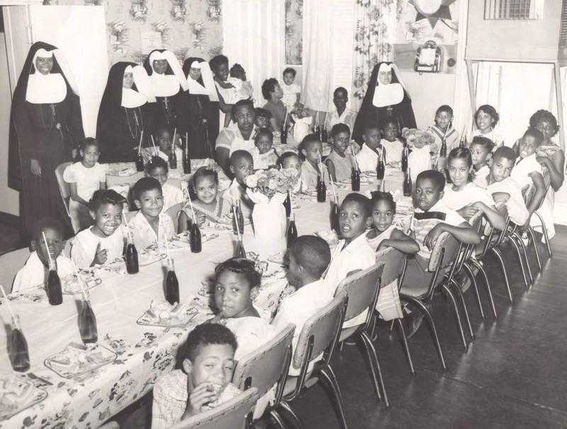 A black and white photo of Oblate Sisters standing behind a long table, full table of children.
