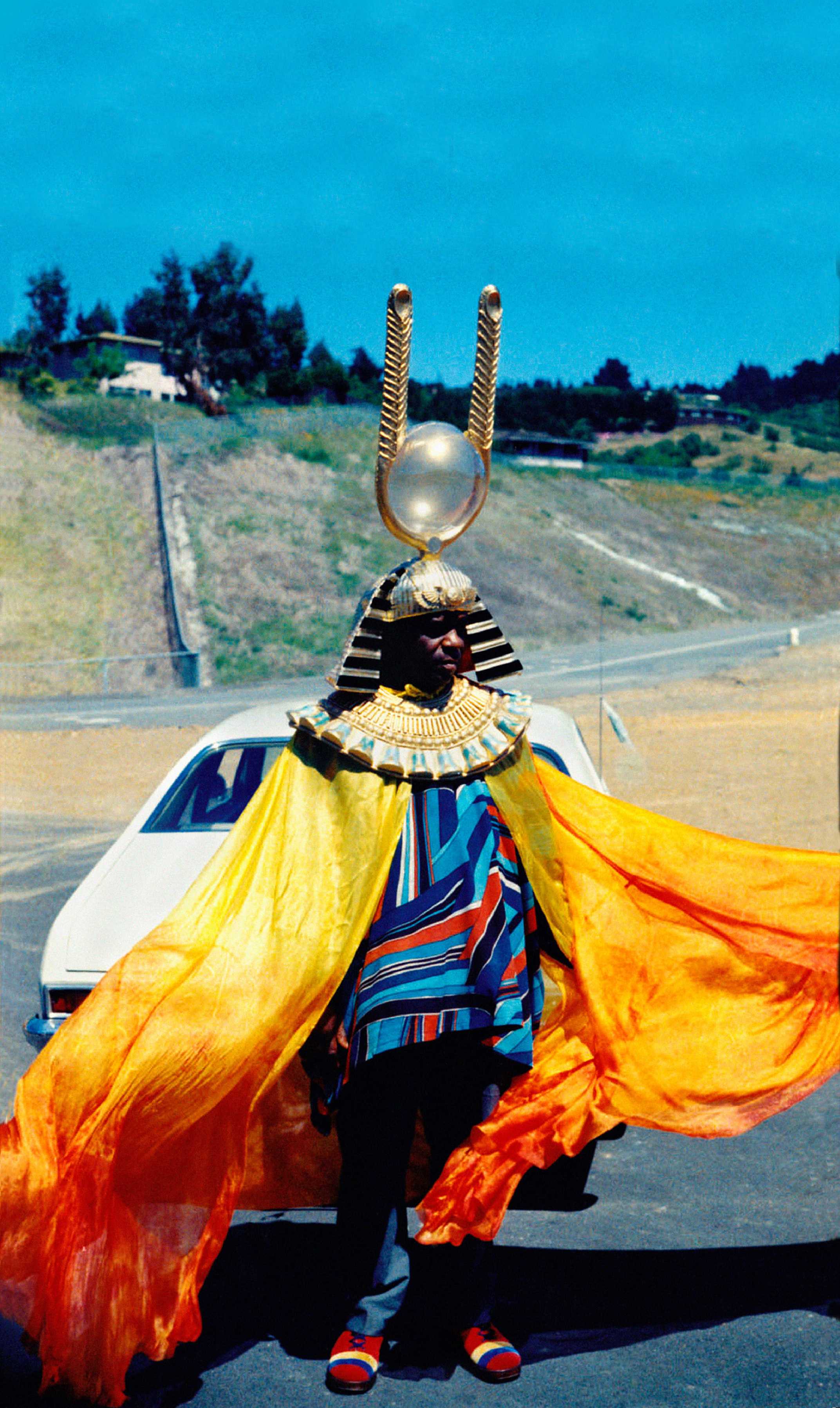 Sun Ra is dressed in vibrant costume complete with a gradient yellow to orange cap and pharaonic headdress.