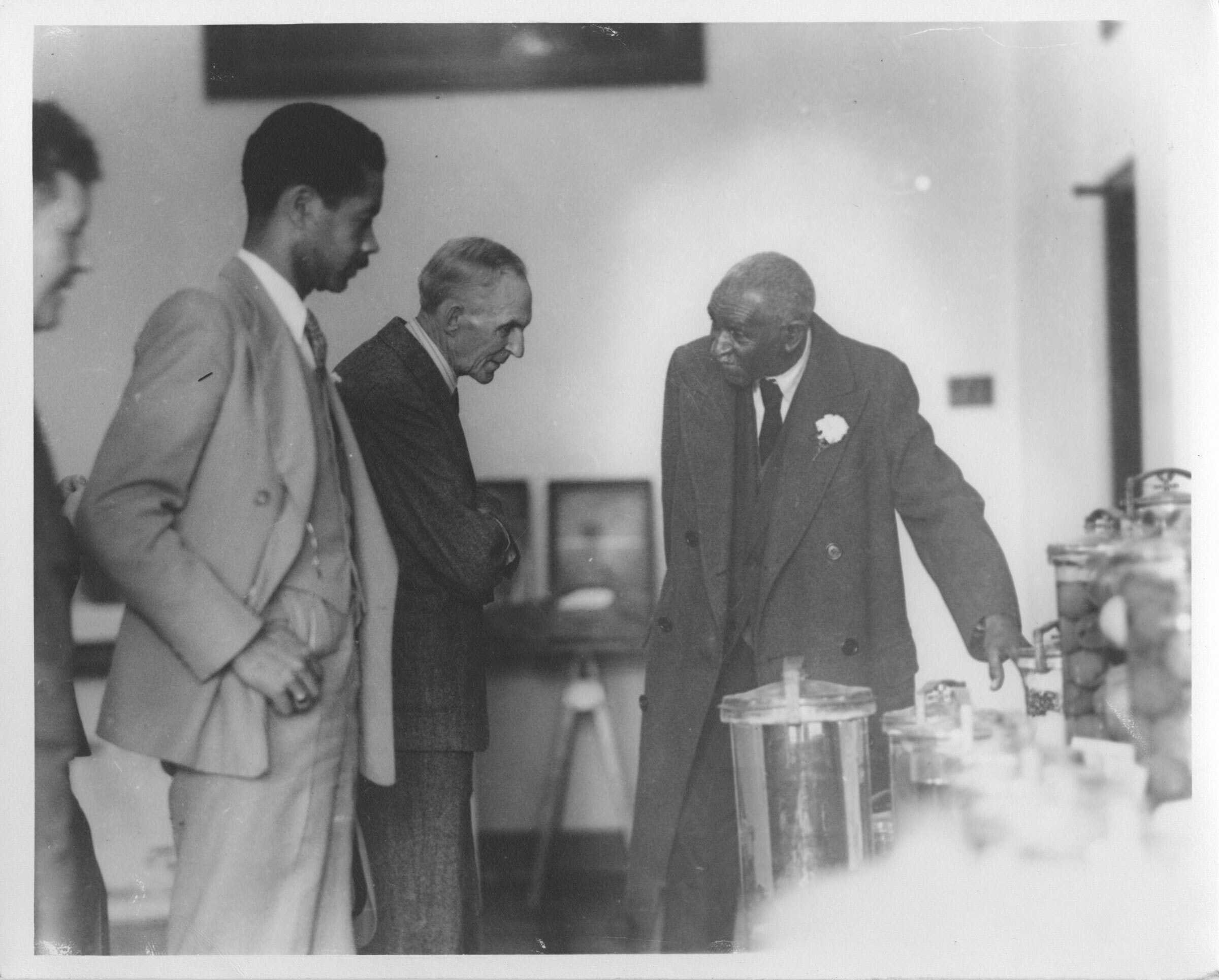 Black and white photograph of George Washington Carver showing Henry Ford his laboratory.  There are two unidentified men looking on.