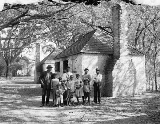 A black and white photograph of a family standing in front of a stone house and a large tree.