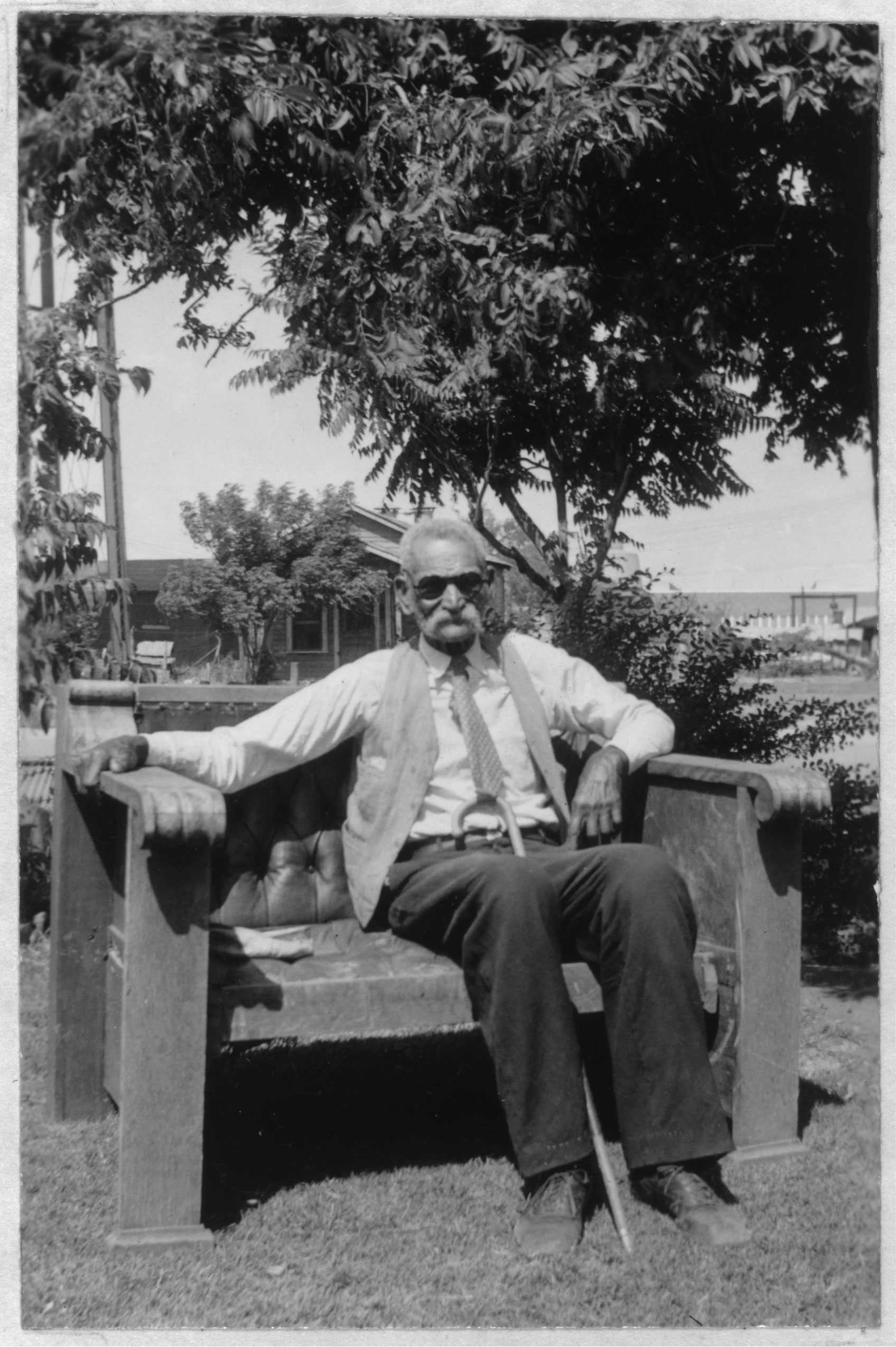 Elderly man wearing sunglasses and a vest sits on a wooden bench in a garden, with trees and a house in the background.