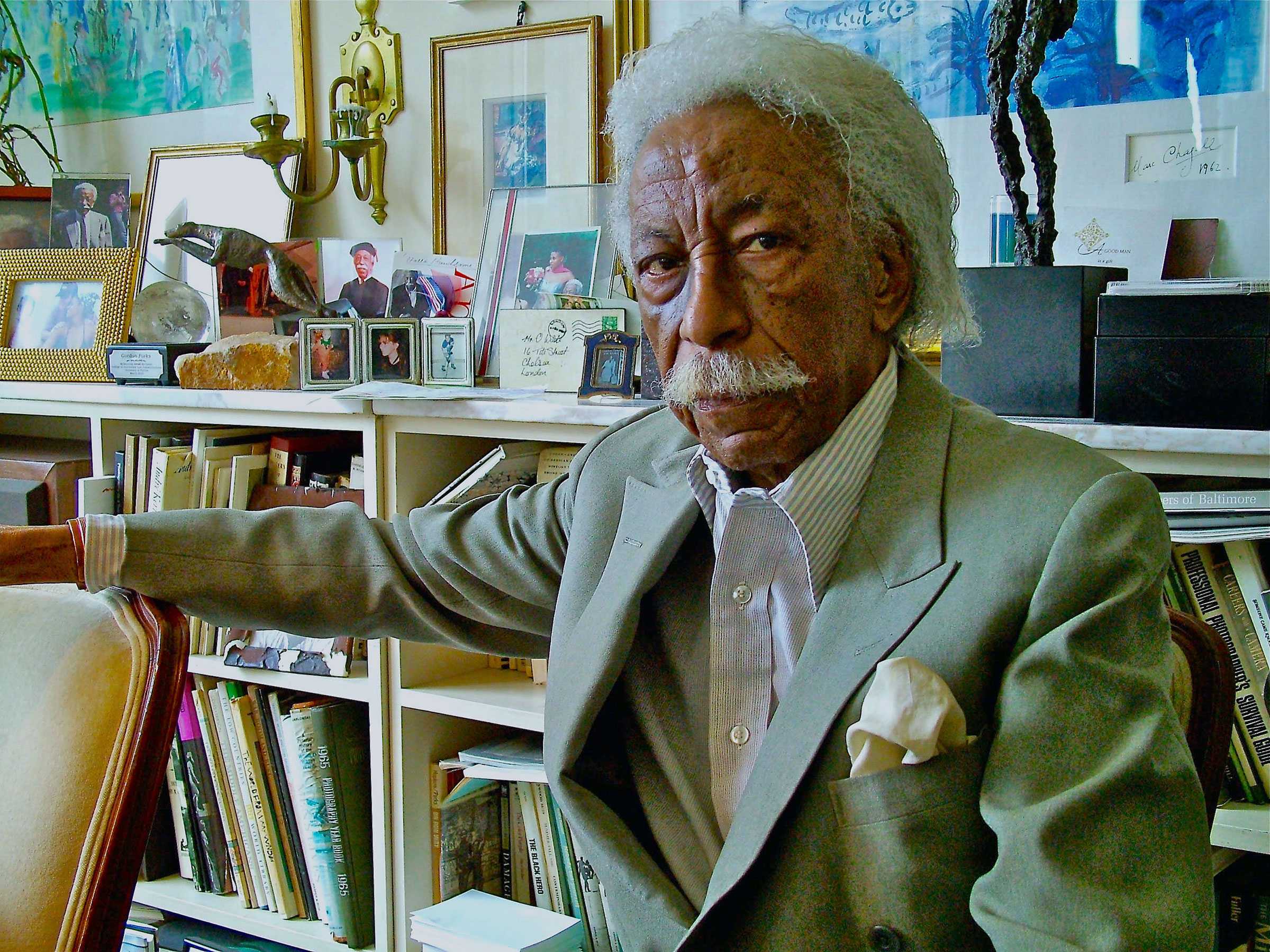 Older man in a suit sitting in a room filled with books and photographs on shelves.