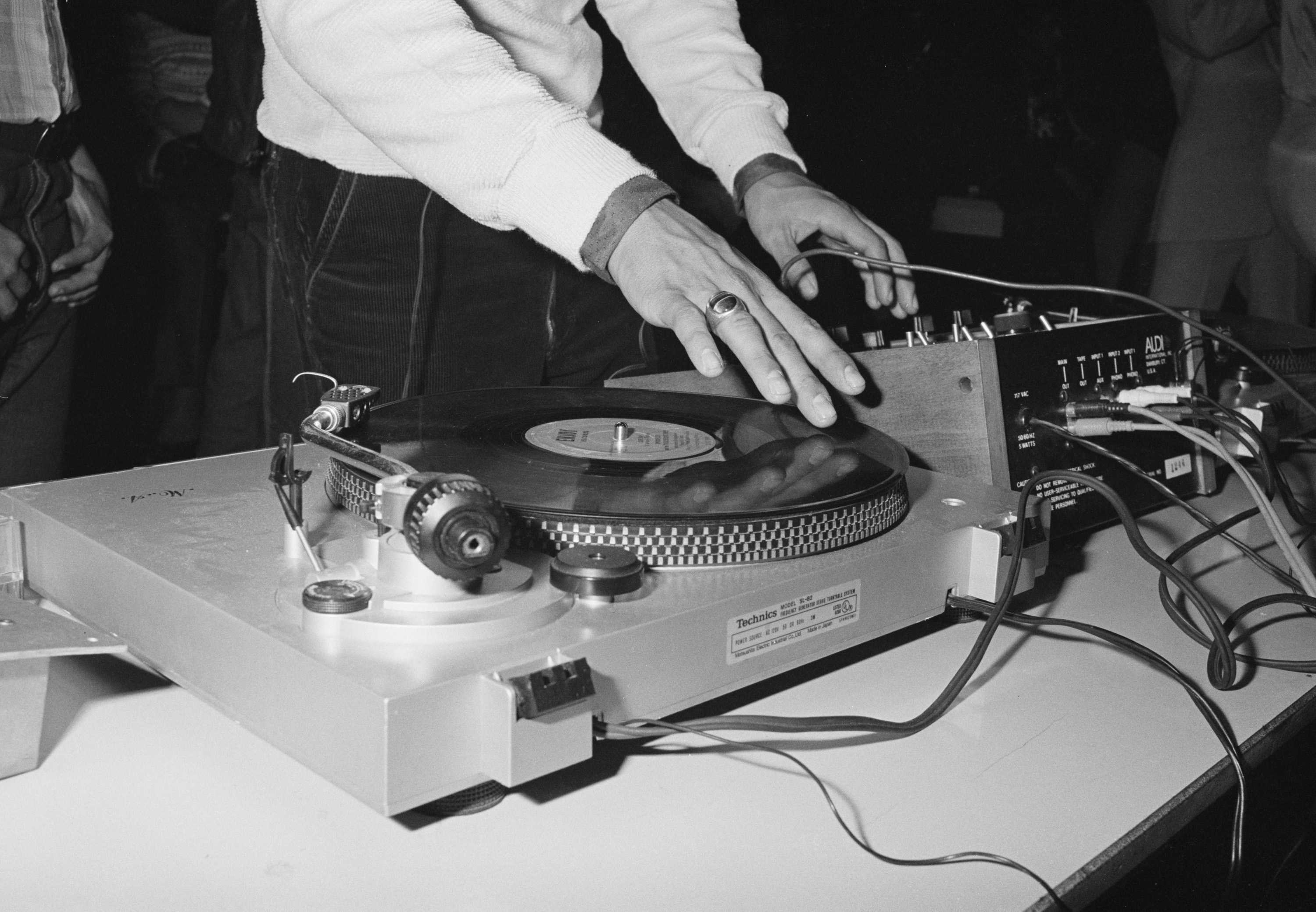 A close up, black and white photo of a someone turning the tables of a record while adjust some gears.