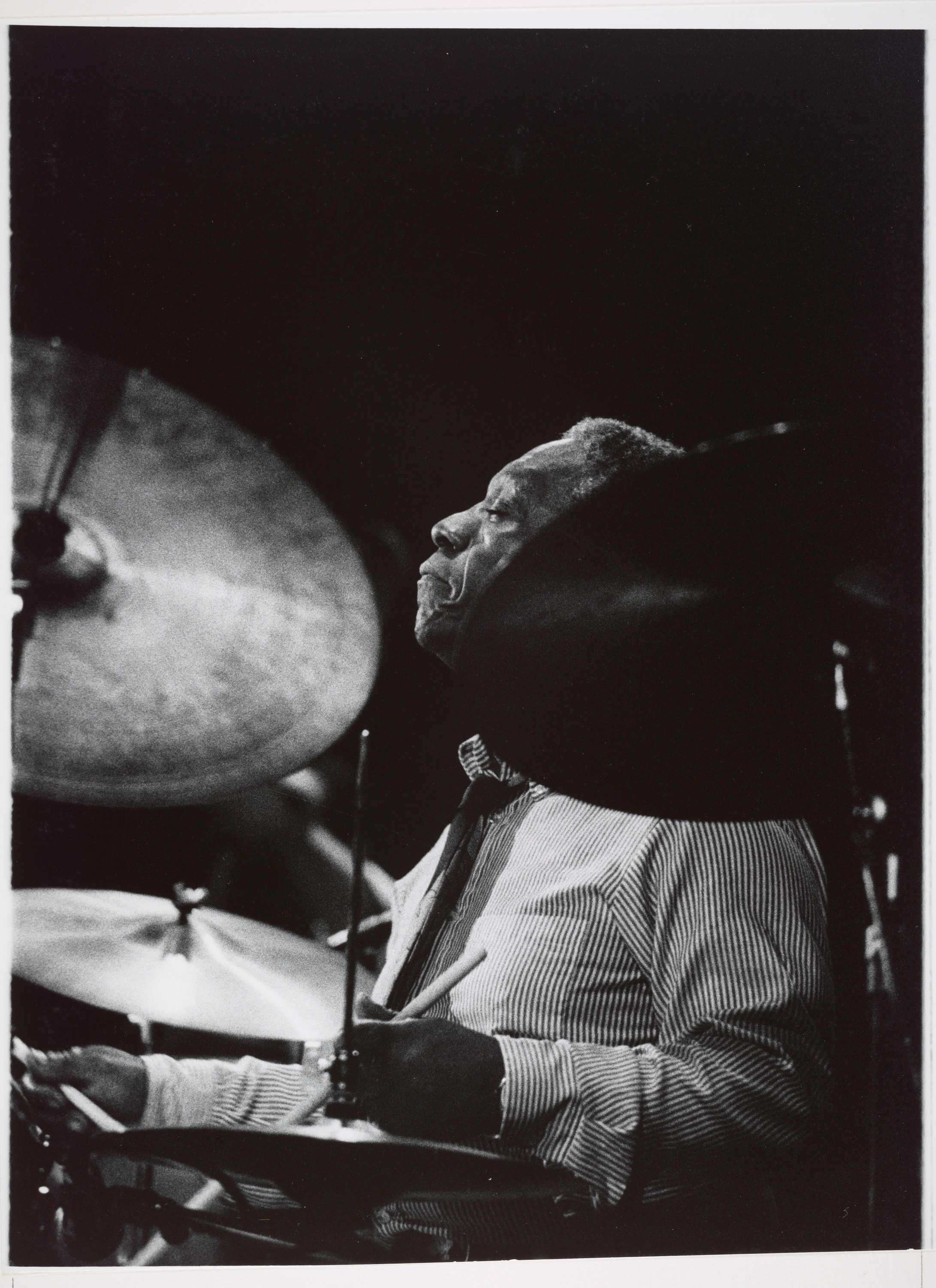 A black-and-white photograph of jazz musician and bandleader Art Blakey playing drums during a 1980 performance. Blakey is photographed in left-profile through his drumkit, with a cymbal partly obscuring his face. His eyes are closed and he wears a striped dress shirt and dark tie.