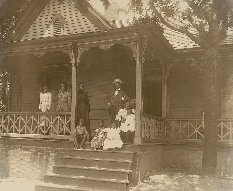 Photograph of unidentified man standing on a porch surrounding by women and children