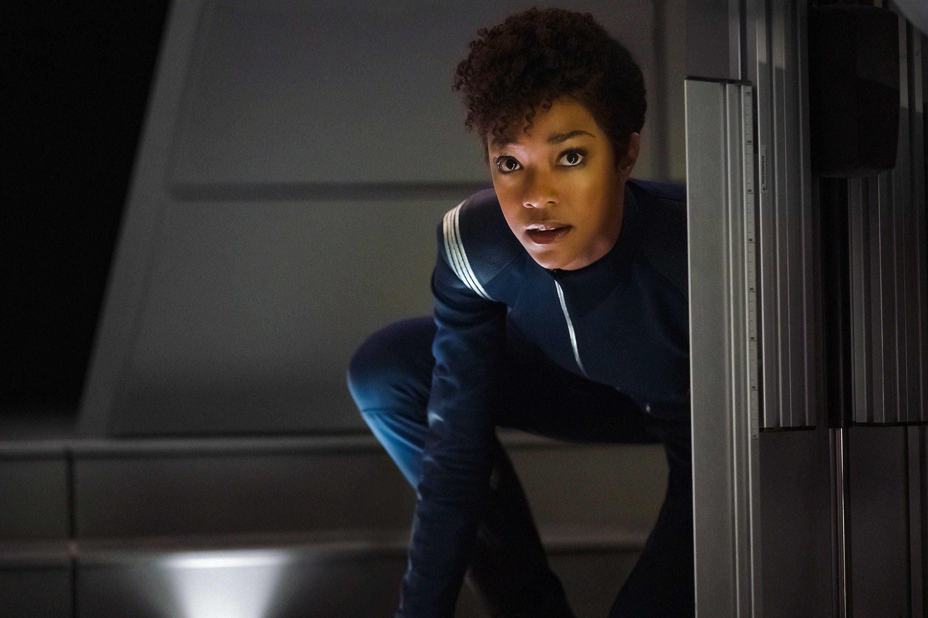 Sonequa Martin-Green is crouched downing, hiding from something. She is wearing a blue space suit.
