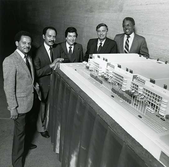Black and white photo of five formally dressed individuals standing behind a detailed architectural model of a building complex. The model includes multiple buildings, flagpoles, and surrounding streets.