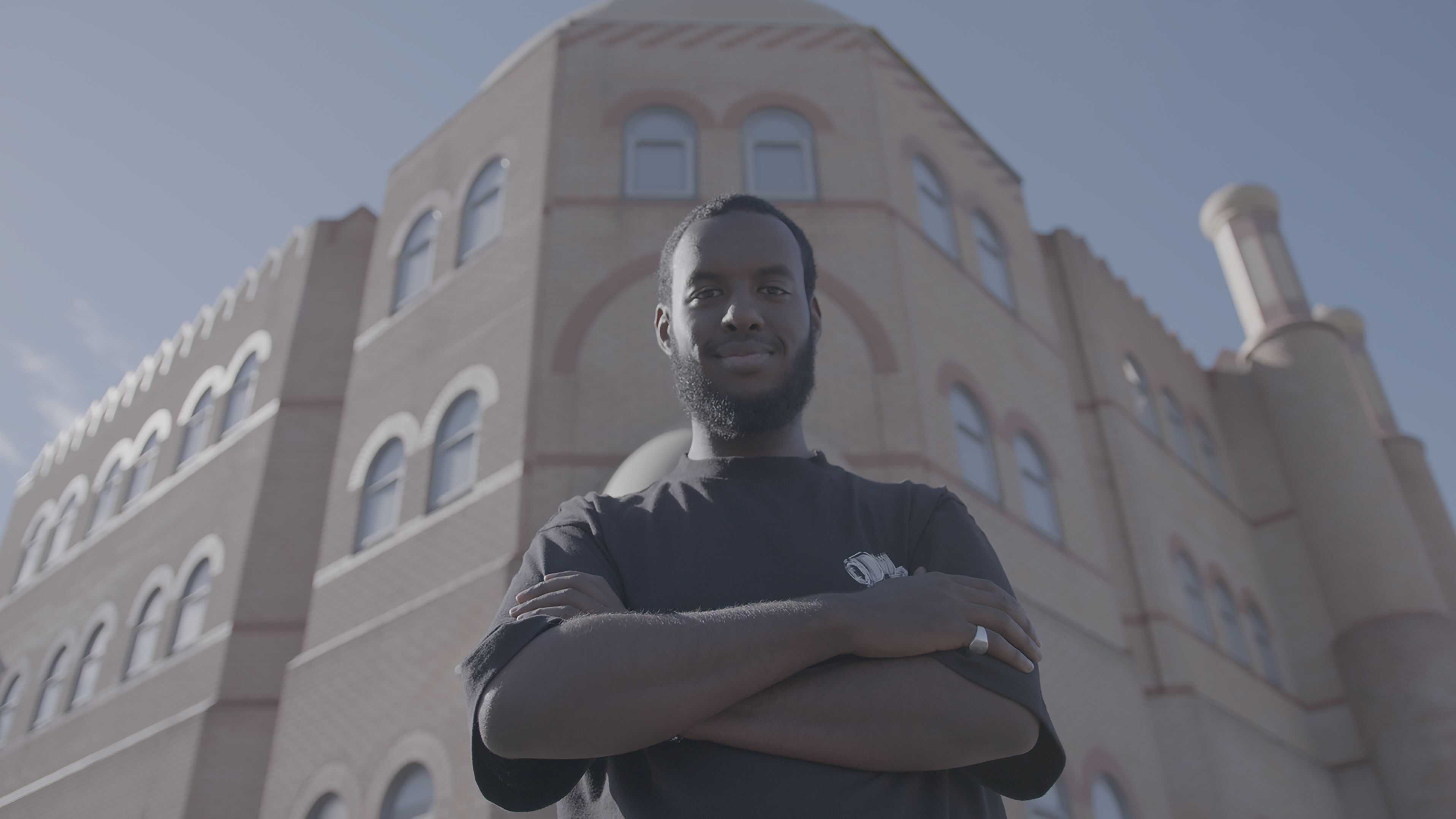 Person standing with arms crossed in front of a large brick building, photographed from a low angle.
