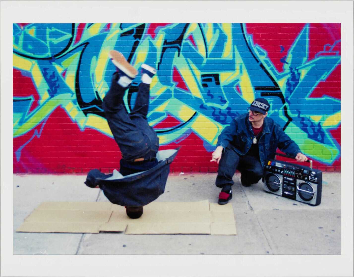 Image of male dancer doing a head spin in front of a mural with another man operating a audio system