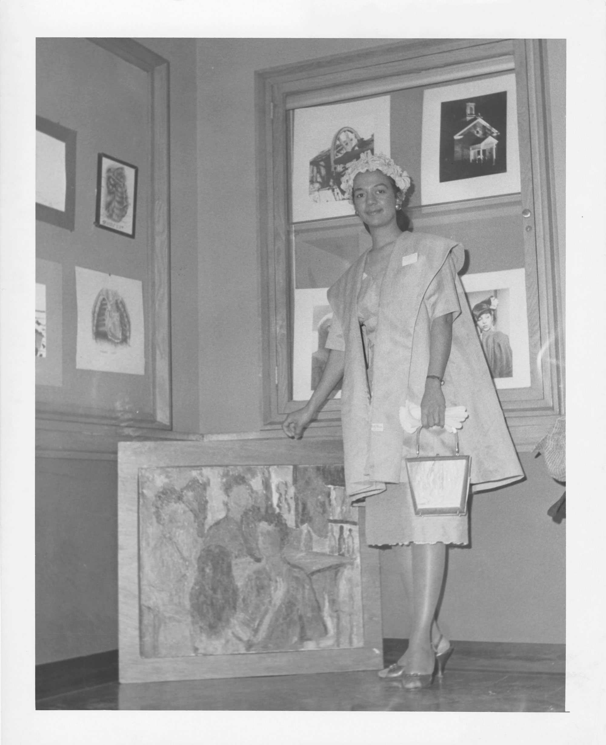 Black-and-white photo of a woman standing in a gallery, with her hand holding up a framed artwork, with more pictures displayed on the wall. behind her.