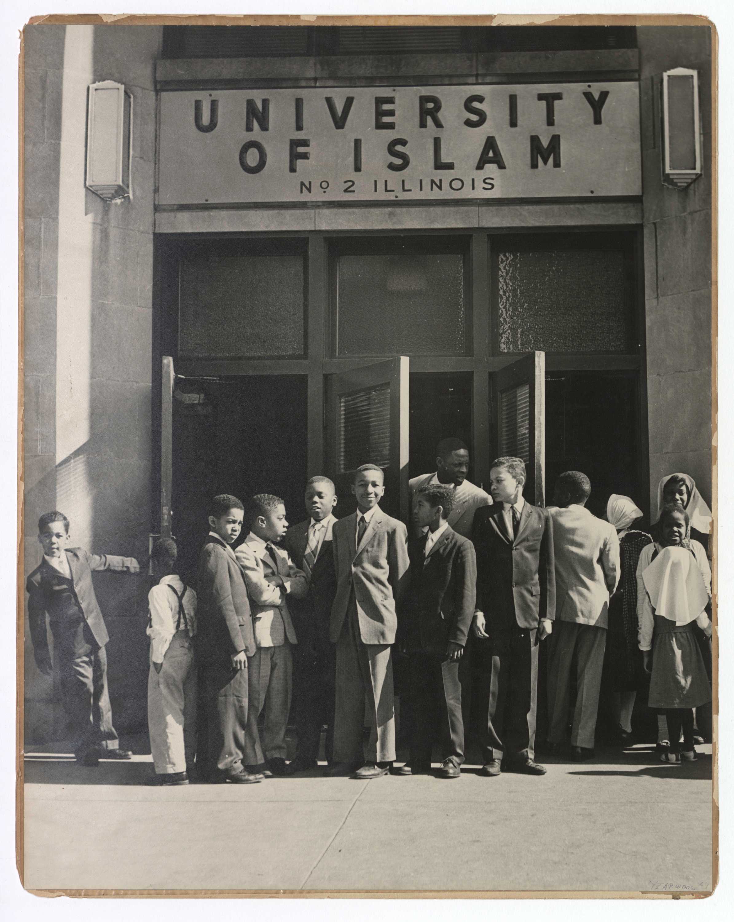 This black and white photograph depicts a group of children standing outside the front doors of the University of Islam.