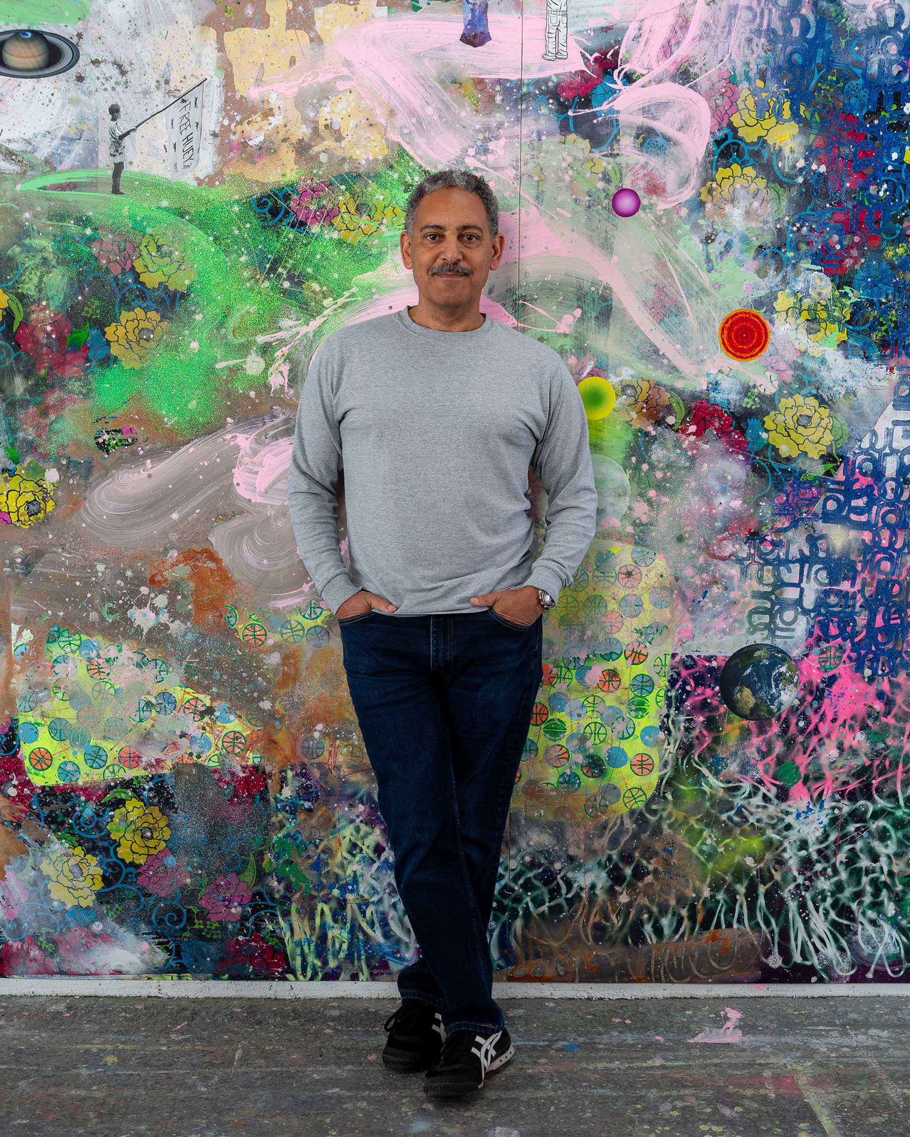 Person standing in front of a colorful abstract painting, wearing a gray shirt and jeans, hands in pockets.