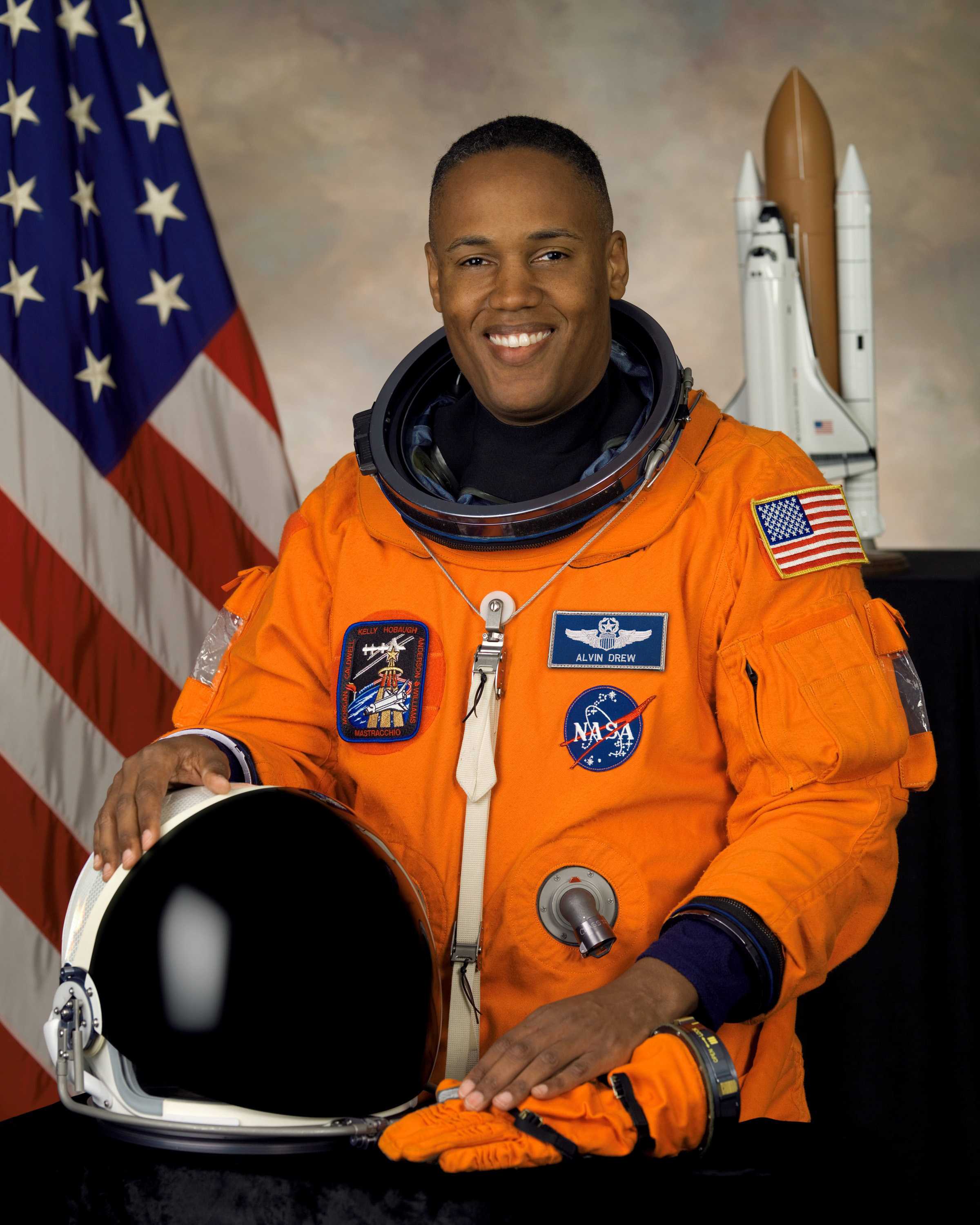 Drew is wearing an orange space suit with a helmet resting on the table in front of him while he smiles for his portrait.