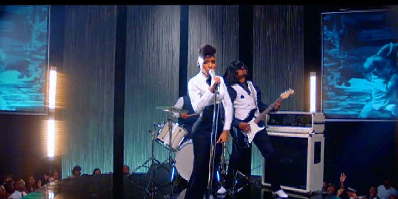 Janelle Monae standing on stage in a white and black suit, singing in front of a crowd.