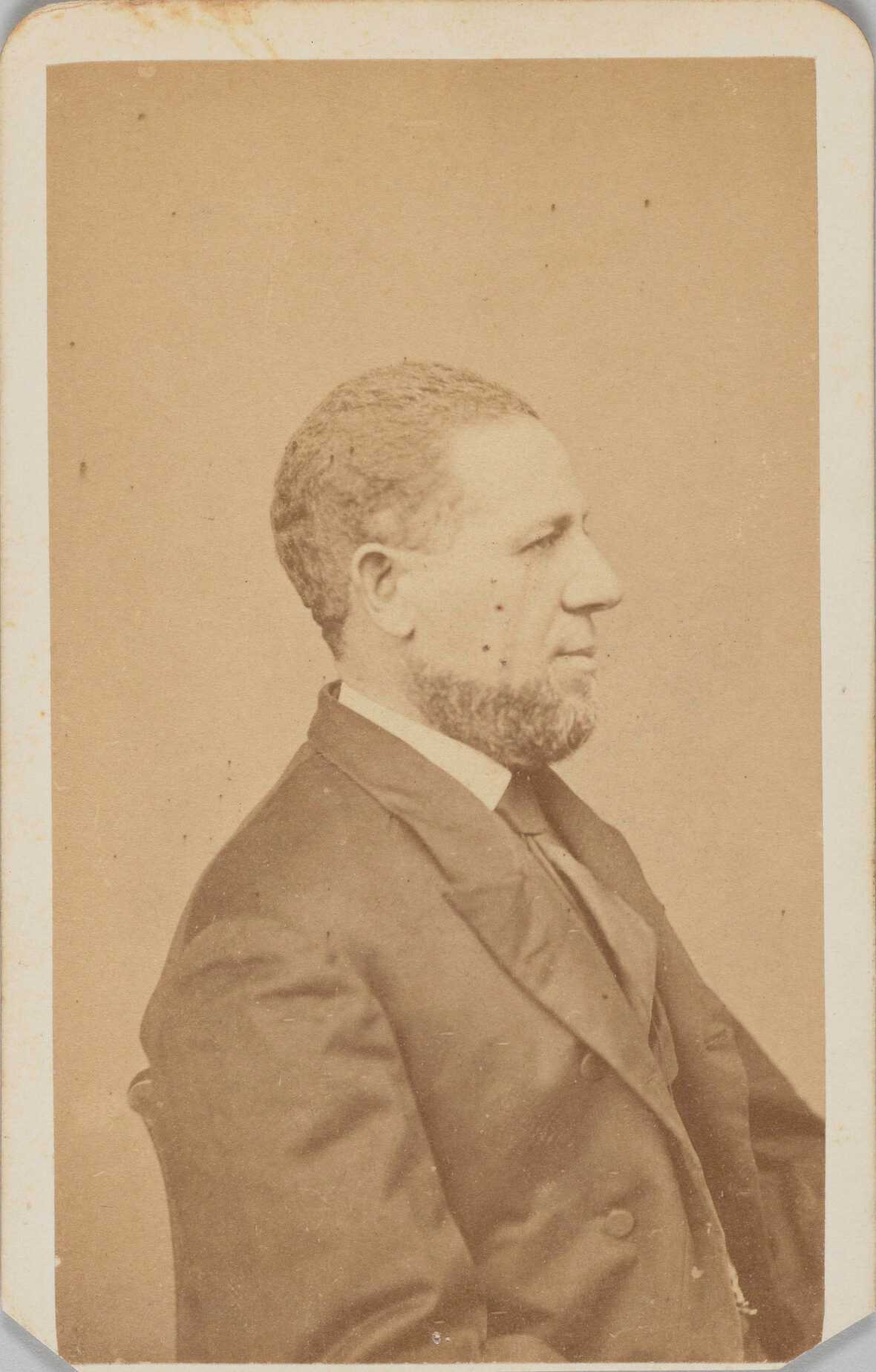 An albumen carte-de-visite portrait of United States Senator Hiram Revels. A dark haired man, Hiram Revels, is seated in a profile position with his proper right side facing the camera. He wears a tall bright collar, dark tie, and dark single breast jacket with two buttons on the torso. The photograph is on original plain mount; the mount is cropped to image along bottom edge. On the back of the photograph, written in pencil, from top to bottom, is: [Hiram Revels / 1st African-American / Senator / Mississippi 1870 / Revells].