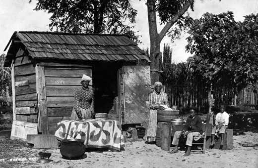 A black and white photograph of two older women washing outside of a shed. Two young boys sit near them.