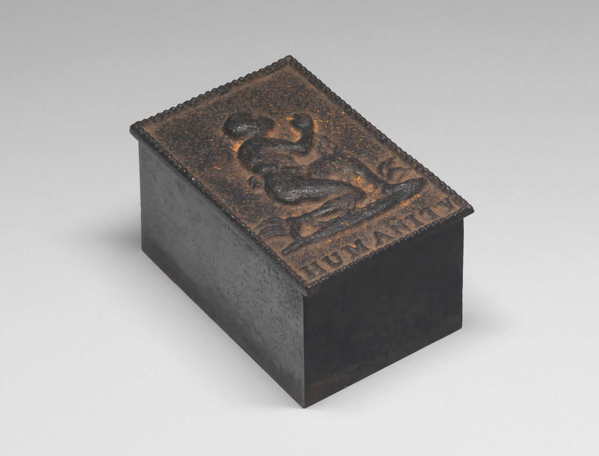 Cast iron desk box, holding a weighted roller blotter. The box bears an embossed representation of a man kneeling in chains, a famous design by Josiah Wedgewood for the English & Foreign Anti-Slavery Society. The single word "HUMANITY" appears below the figure and a raised, decorative border is around the edge. The box contains a brass topped iron piece with a slightly curved bottom.