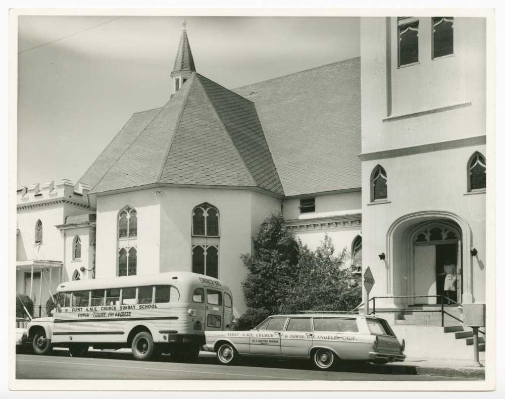 Black and white photograph of a church as seen from the street. The church has a white exterior with gothic style windows of stained glass. On the left half of the image is a multi-walled transept extending from the building. Above this in the background is a cupola. In the foreground in the street on in the bottom left corner is a bus with [FIRST A.M.E. CHURCH SUNDAY SCHOOL/EIGHTH and TOWNE, LOS ANGELES] painted on the side. Behind the bus, right of center, is a sedan with [FIRST A.M.E. CHURCH 8th & TOWNE, LOS ANGELES/DR. H. HARTFORD BROOKINS/Minister] painted on the side. In the lower right corner of the image is an arched and covered doorway with a woman in white emerging. On the reverse of the image is a stamp in the center set at a diagonal [J. BANKS/HOME PORTRAITURE PHOTOGRAPHER].