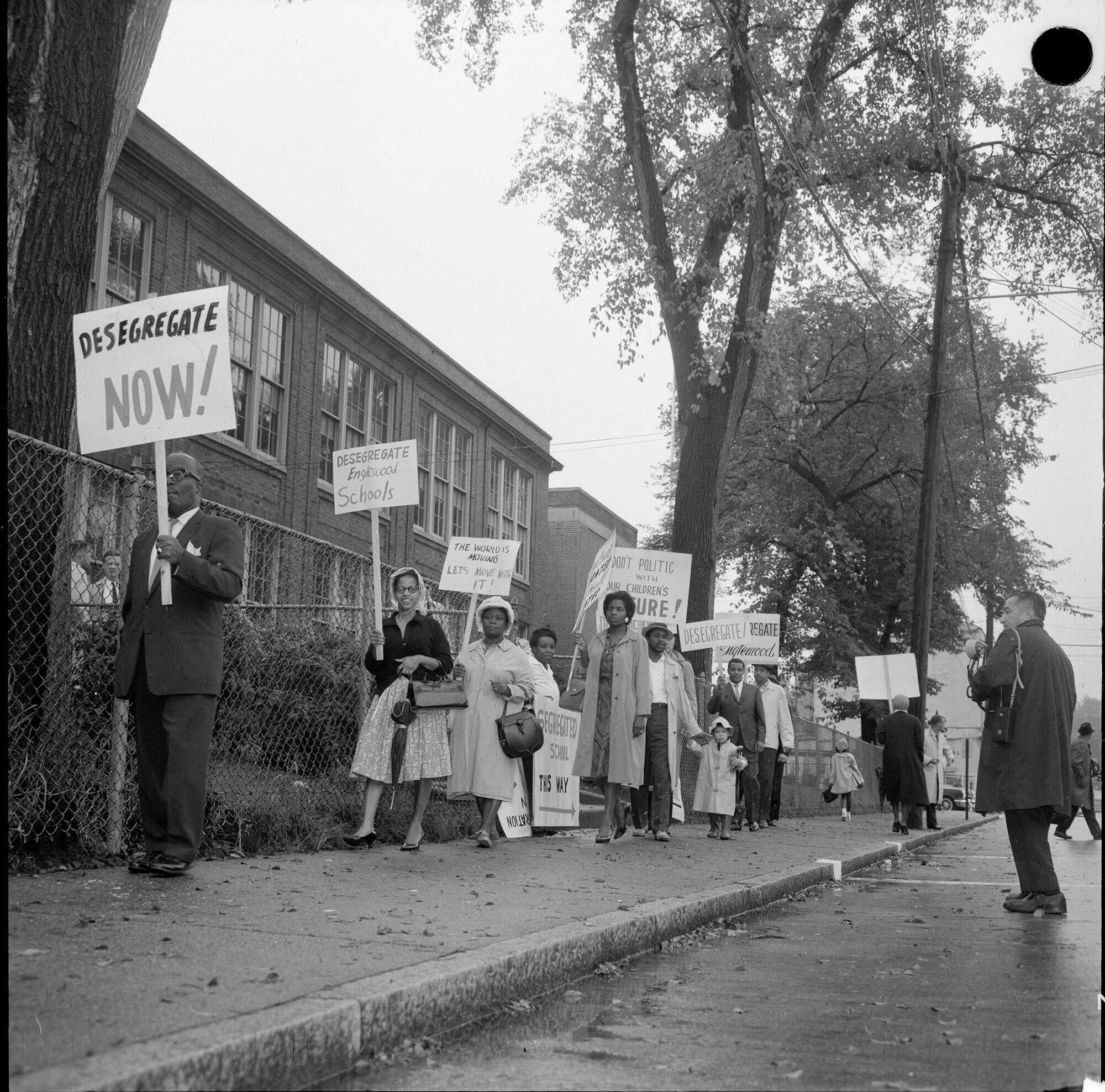 Black and white photograph of people protesting holding signs that say "Desegregate Now".