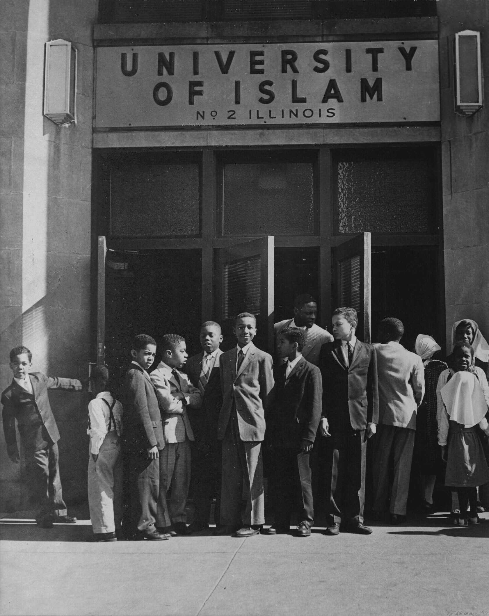 This black and white photograph depicts a group of children standing outside the front doors of the University of Islam.