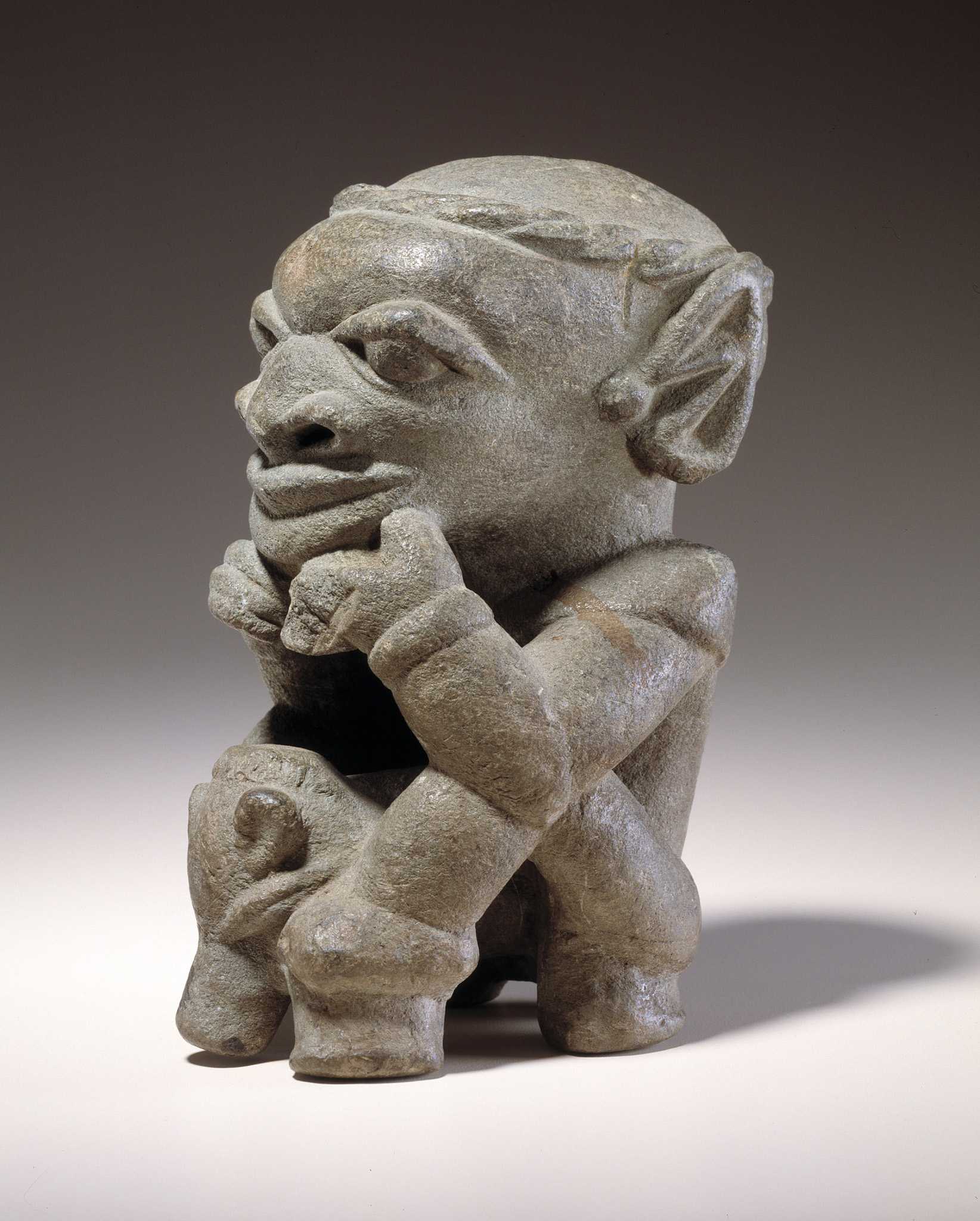 A stone figurine of Nomoli sitting with its knees bend and resting its head on its hands.